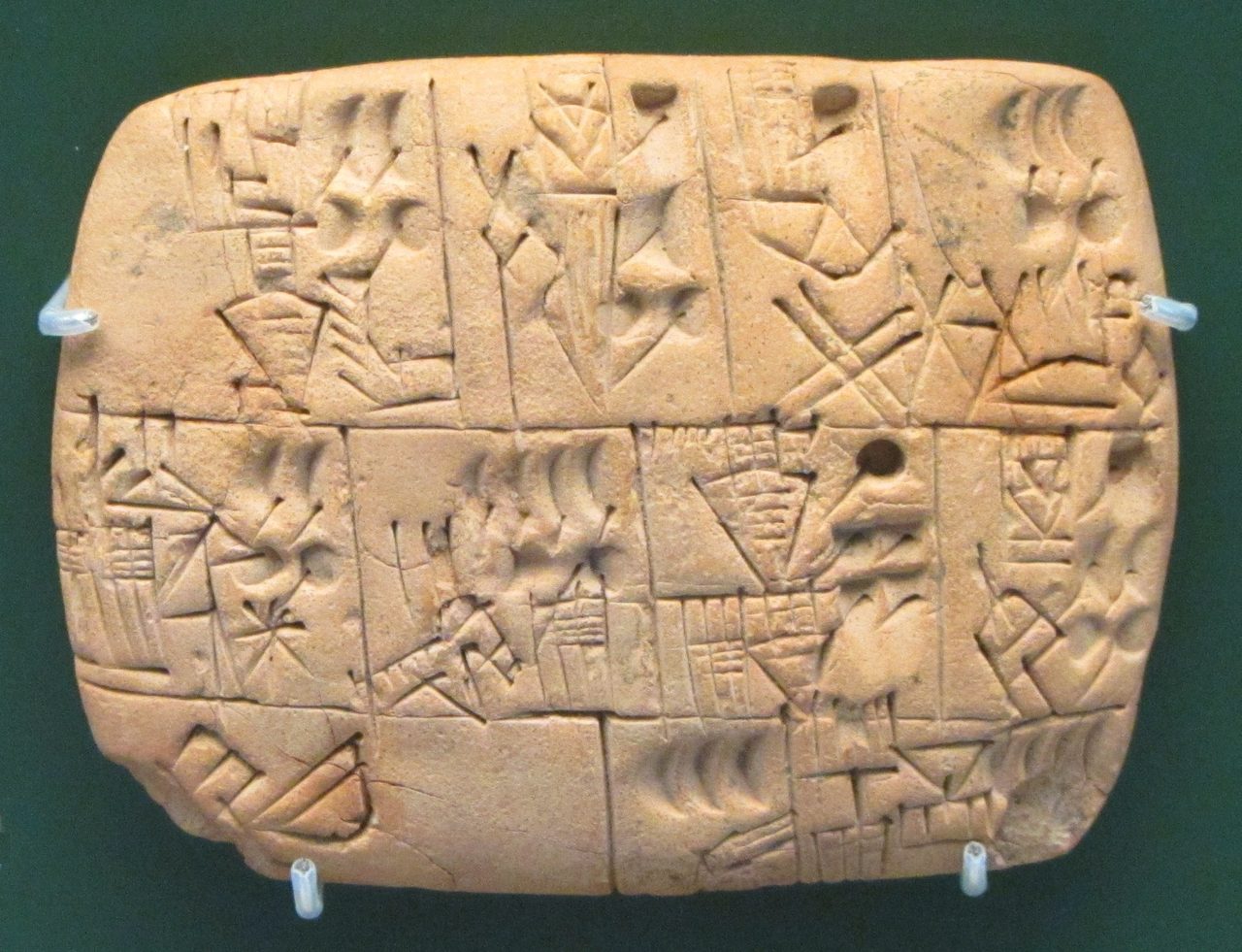 A small pale clay tablet photographed against a plain background, its surface ruled into a grid of rectangular cells carrying small impressed marks: rounded punches and finer incised signs.