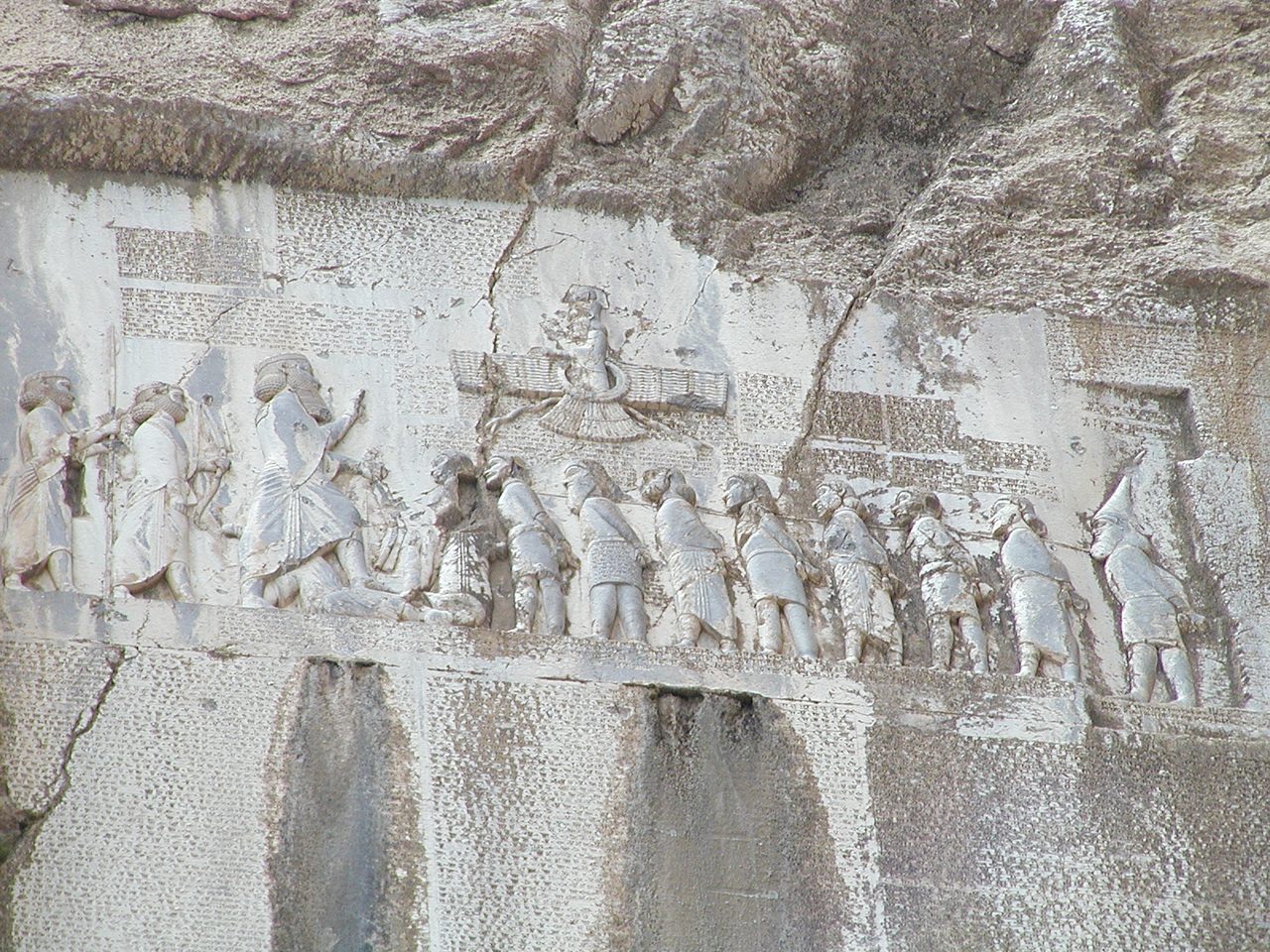 A photograph of a rock face on a mountainside: a carved relief panel of standing figures, with large rectangular blocks of columnar inscription cut into the cliff beside and below it.