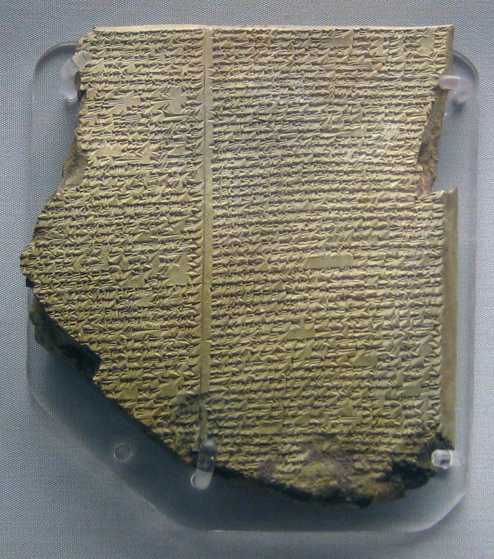 A broken clay tablet densely covered in small cuneiform signs. Its top edge is an intact original rim, with the lines of text ending cleanly against it at both corners; the left side, the bottom and the whole lower-left corner along a long diagonal are lost, and a strip is missing from the right edge below the top corner.
