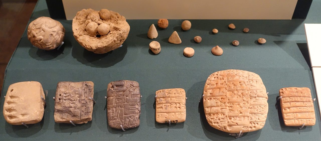 A wide photograph of a lit museum display case holding small clay objects on a plain mount: a hollow clay ball, a group of small geometric counters, and several inscribed clay tablets.