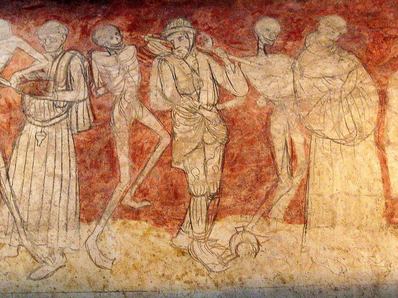 A weathered wall fresco in earth reds and ochres, the figures drawn in thin brown outline: a robed figure holding a book at the left, a shrunken corpse figure leaning toward a man in a belted tunic and hose who carries a long handled tool, a second corpse laying a hand on that man's shoulder, and a figure in a long cowled robe at the right