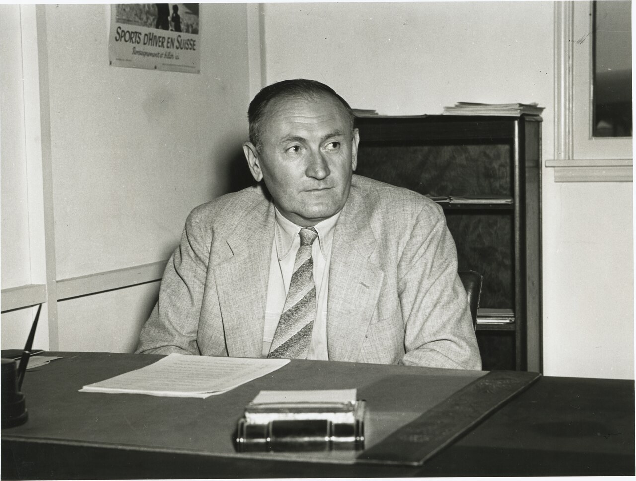 A black-and-white photograph of Fritz Zwicky as a middle-aged man in a light suit and striped tie, seated at a desk in an office, a winter-sports poster on the wall behind him