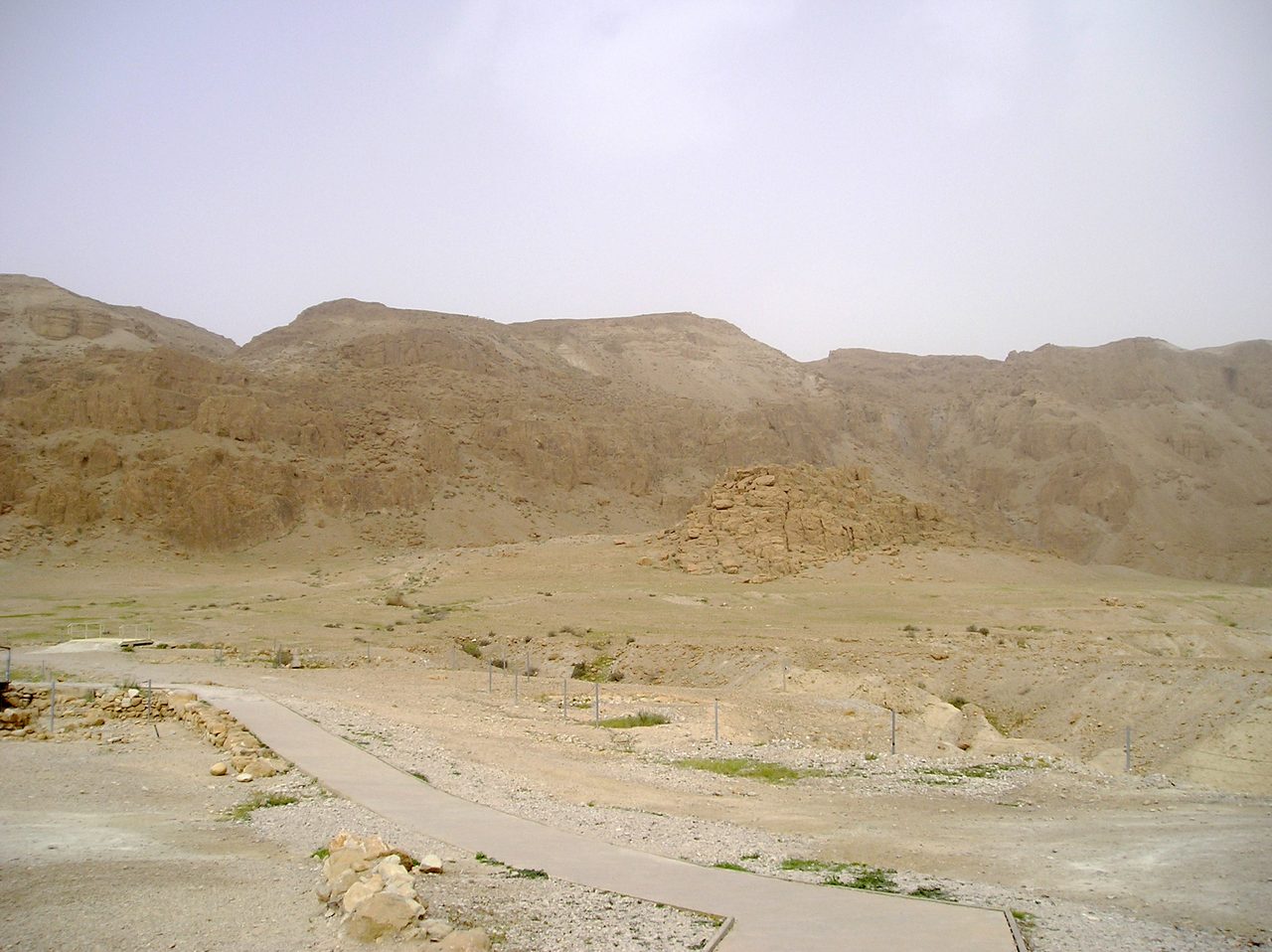 An eroded pale marl and limestone slope in the cliffs at Khirbet Qumran, with a modern paved walkway and metal fence posts crossing the foreground