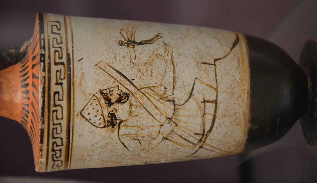 An ancient Greek white-ground lekythos (funerary vase) shown on its side, painted in ochre line with Charon the ferryman: a bearded figure in a pointed cap leaning on his long pole, with a Greek-key border running along the vase's shoulder and its black neck and foot at either end
