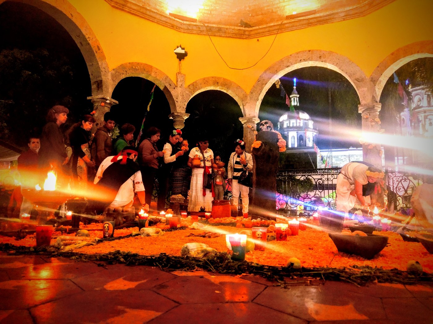 A nighttime Day of the Dead (Dia de los Muertos) gathering in a yellow arched Mexican courtyard: people in traditional white dress stand around a large floor offering of marigold petals, dozens of lit candles, bowls, and a skull, with rays of light crossing the scene