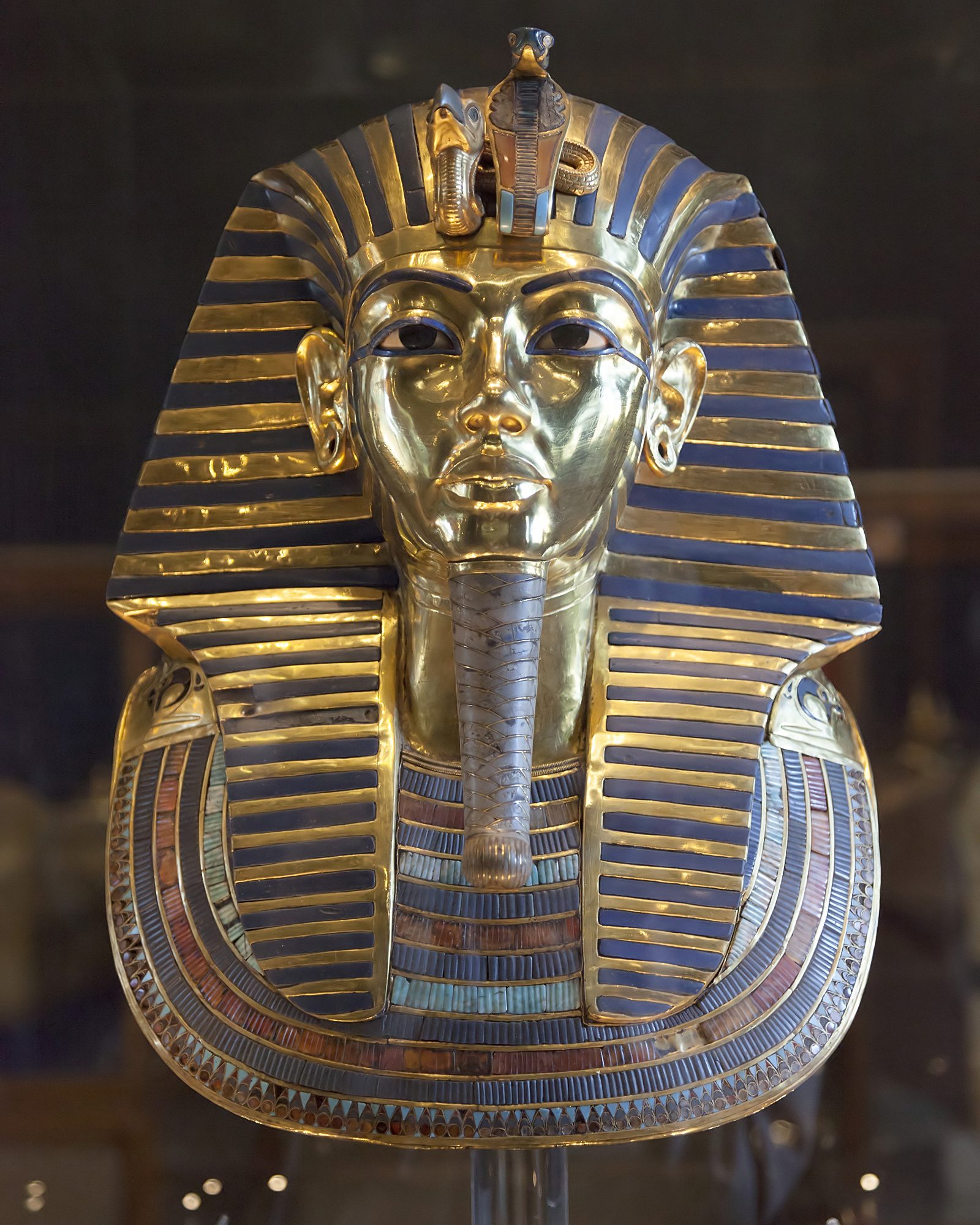 The gold funerary mask of Tutankhamun on a stand against a dark ground. A smooth polished gold face with dark inlaid eyes and blue inlaid brows and eyeliner, under a striped headdress of alternating gold and deep blue bands falling to the shoulders. A vulture head and a rearing cobra project from the brow. A long narrow plaited beard hangs from the chin, and a wide collar of inlaid coloured stone lies across the chest.