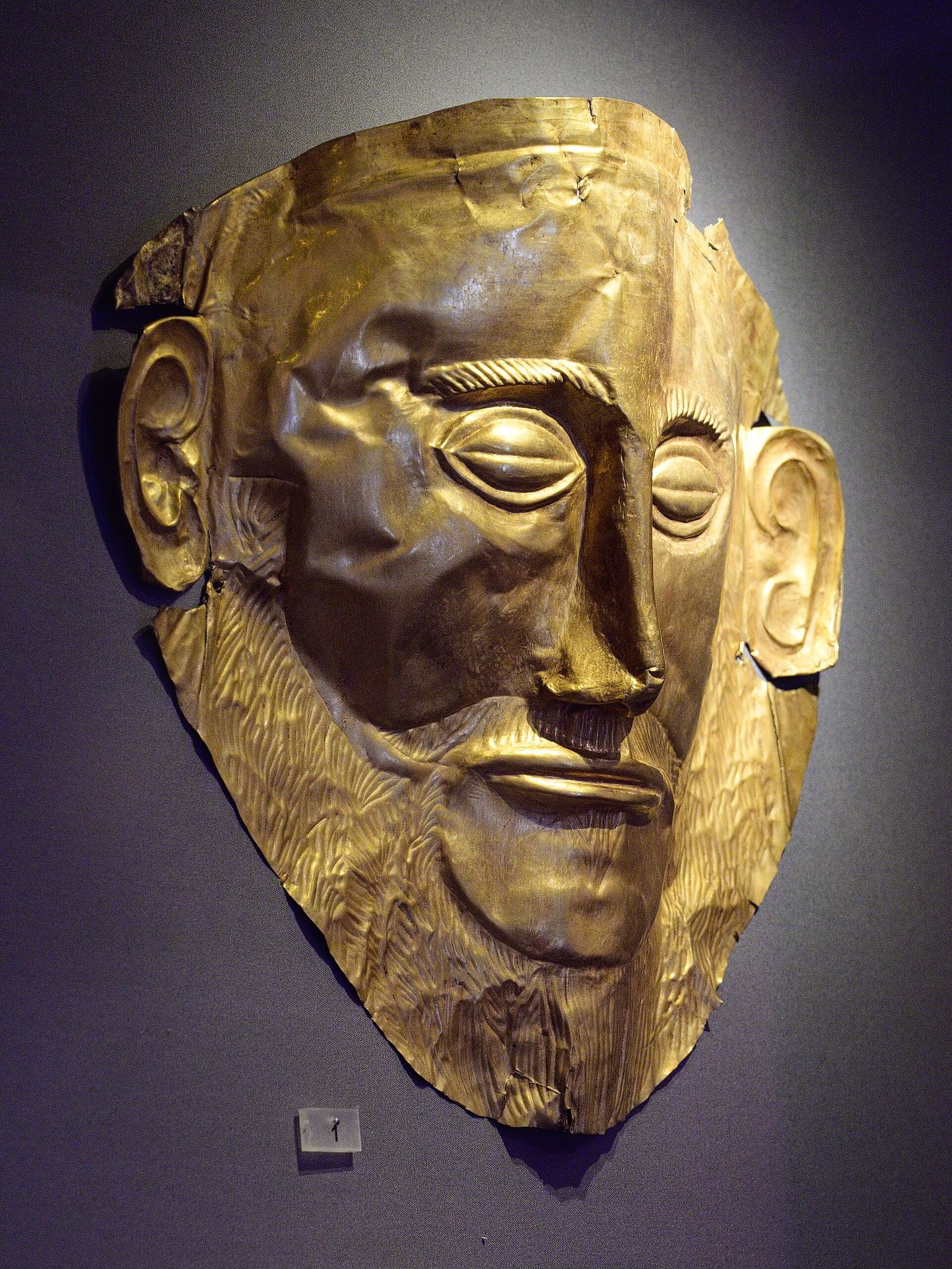 A gold mask mounted on a grey wall and lit from above. The face is long and narrow, with closed lidded eyes, a straight prominent nose, a moustache and a pointed beard rendered in fine parallel chased lines, and a thin closed mouth. The ears stand clear of the surrounding sheet, cut free of the body of the mask. The gold is buckled and torn along the lower and left edges.