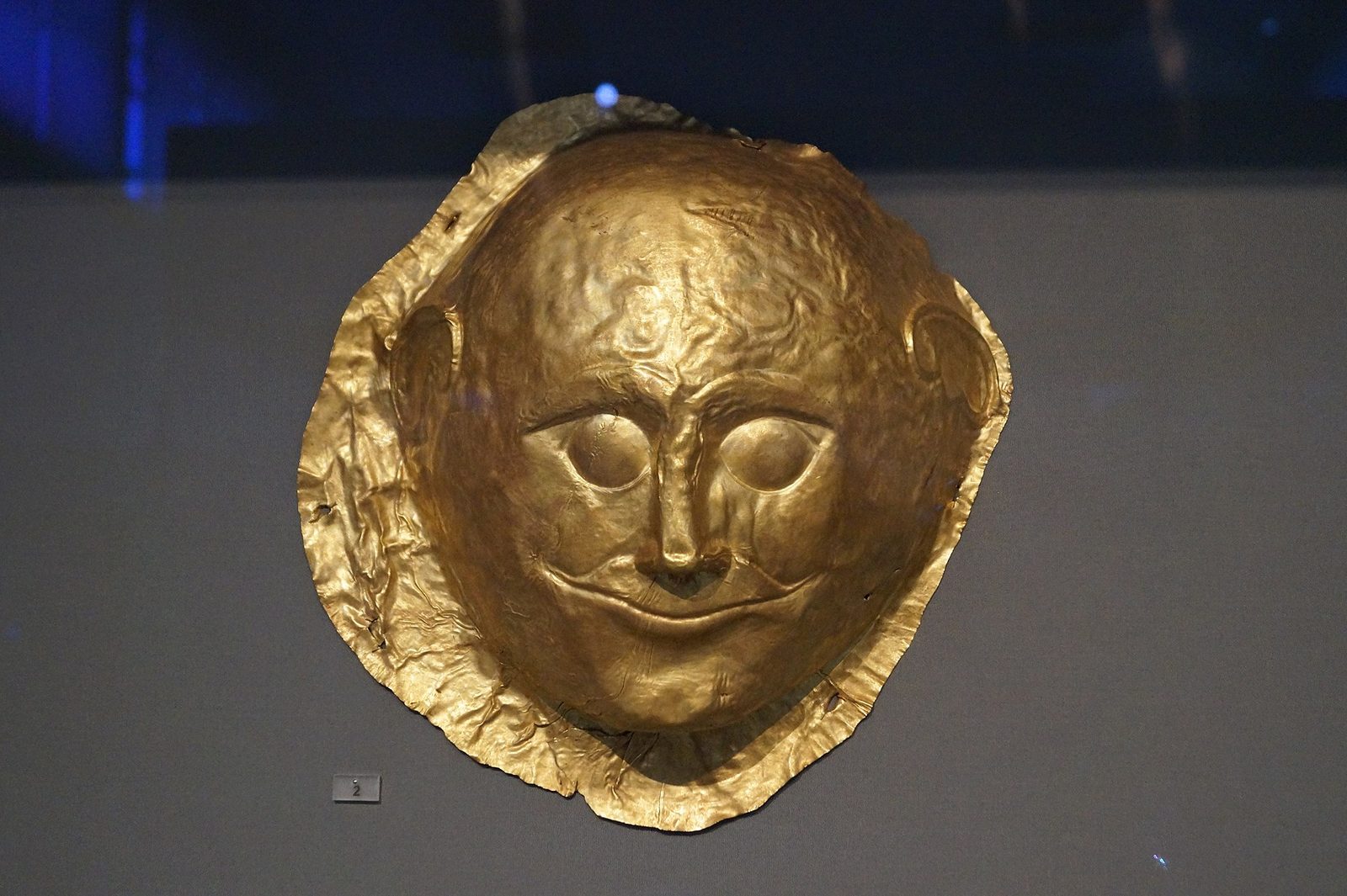 A gold mask lit against a dark museum ground. The face is broad and round, with closed almond eyes, a small straight mouth turned faintly up at the corners, and ears worked into the metal at each side. The gold is thin sheet, buckled and creased around the rim where it has been shaped, and the whole object reads as a flattened oval a little larger than a face.