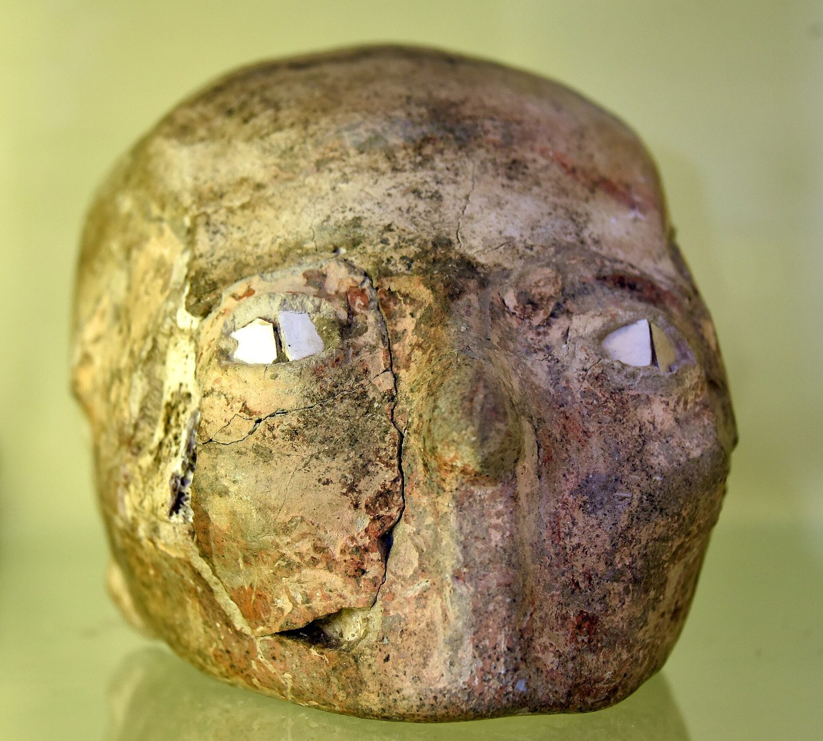 A human skull covered in ochre and cream plaster, photographed close against a pale green ground. The plaster is built up over the bone into a rounded face with a low brow and a broad flattened nose. Two irregular pieces of white shell are set into each eye socket in place of eyes. Cracks run down the length of the face and across the crown, and the lower jaw is absent.