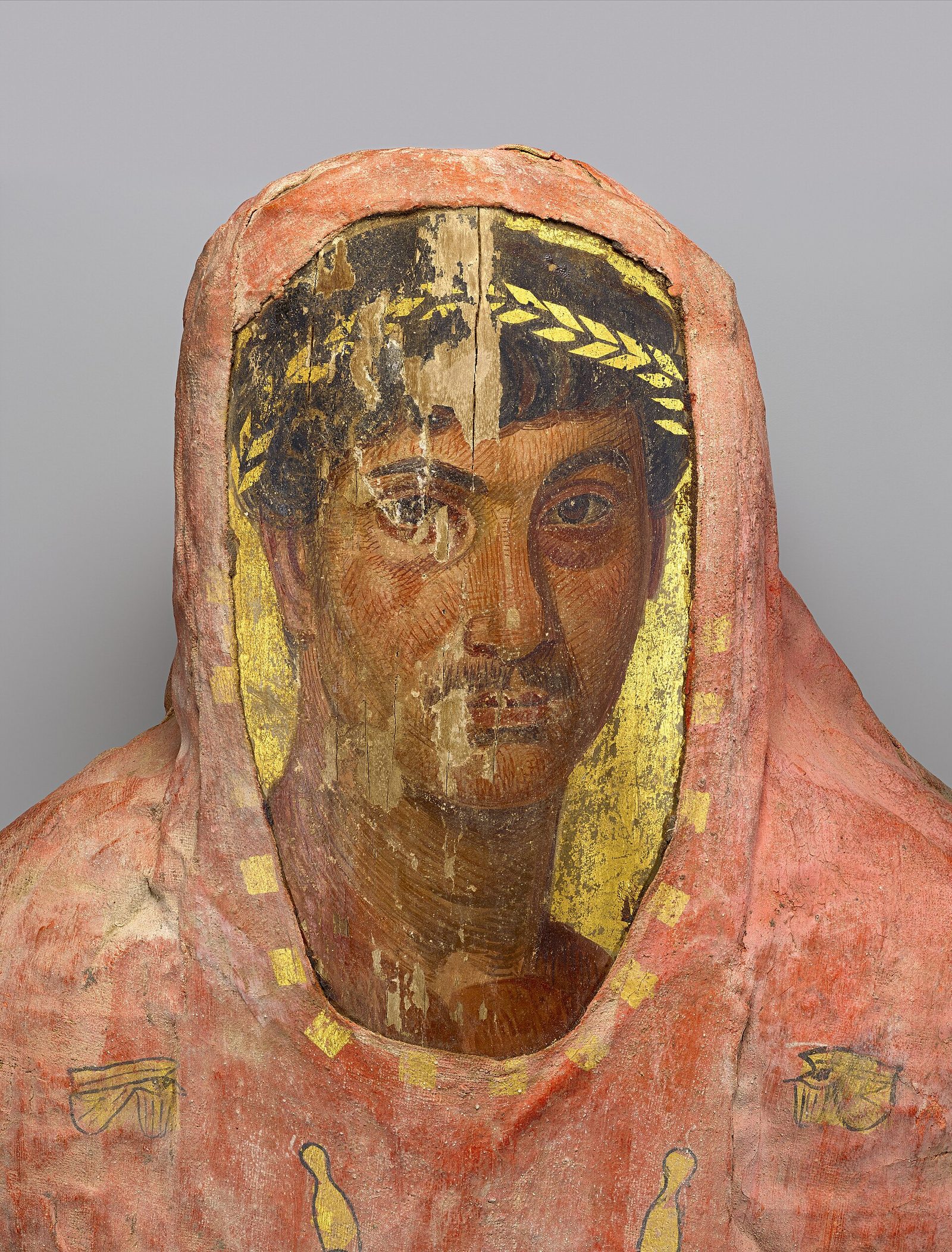 The head and shoulders of a wrapped mummy, its linen covered in dull pink-red plaster with small gilded marks. A rounded opening is cut in the wrappings over the face, and set into it is a flat wooden panel painted with the portrait of a young man: dark curly hair under a gilded laurel wreath, large dark eyes, a faint moustache. Gold leaf fills the space between the painted panel and the edge of the opening. A vertical split runs down the middle of the panel and the wood grain is visible through the paint.