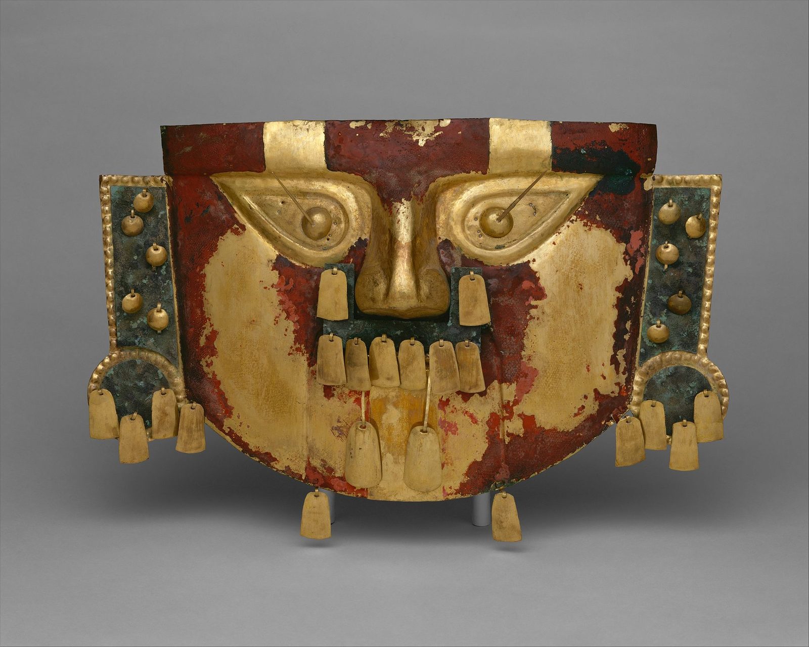 A broad, flat gold mask, wider than it is tall, on a grey ground. Large almond eyes with raised centres slant upward at the outer corners, each with a thin pointed projection running out toward the temple. The nose stands out in relief. Much of the gold surface is stained a deep brick red, heaviest across the brow and the sides of the face. Rectangular panels of green-corroded metal are riveted at each side, and rows of small gold bangles hang from the mouth area and from the lower edges.
