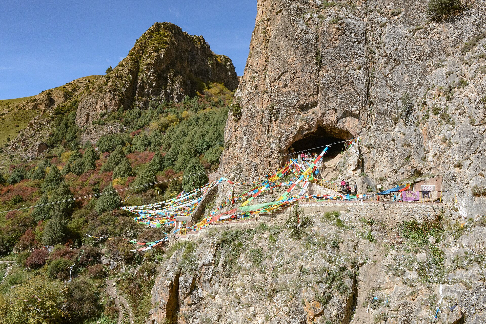 A pale limestone cliff on the right with a dark arched cave mouth partway up it. Strings of Tibetan Buddhist prayer flags in blue, white, red, green and yellow radiate from the cave mouth down and to the left across a gorge on cables. A stone-walled path and terrace run along the cliff base below the cave, with small figures near the entrance and on the path and a small shrine at the right. Across the gorge a rocky ridge carries green scrub and autumn-coloured trees under a clear blue sky.