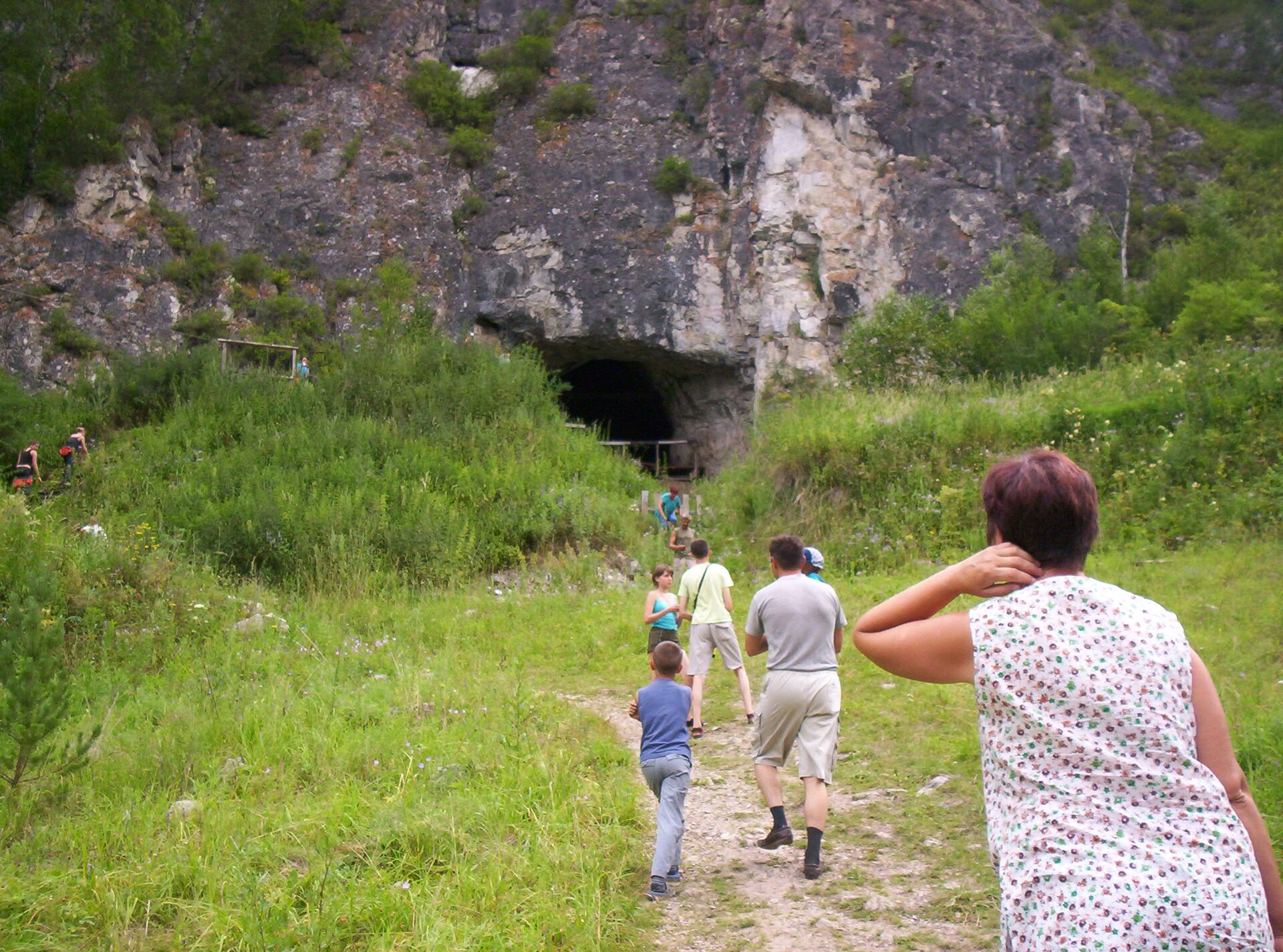 A casual outdoor snapshot. Tourists walk up a worn grass-and-gravel path toward the dark arched mouth of a cave in a grey limestone cliff, under summer greenery. A family group is on the path, more figures stand near the cave mouth, and a wooden platform is visible on the slope to its left. A woman in a floral sleeveless top stands in the right foreground with her back to the camera.