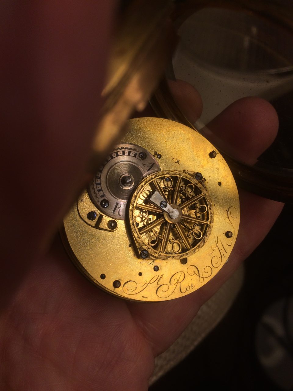 A hand holding an opened pocket watch, its case swung back to show the back plate of the movement