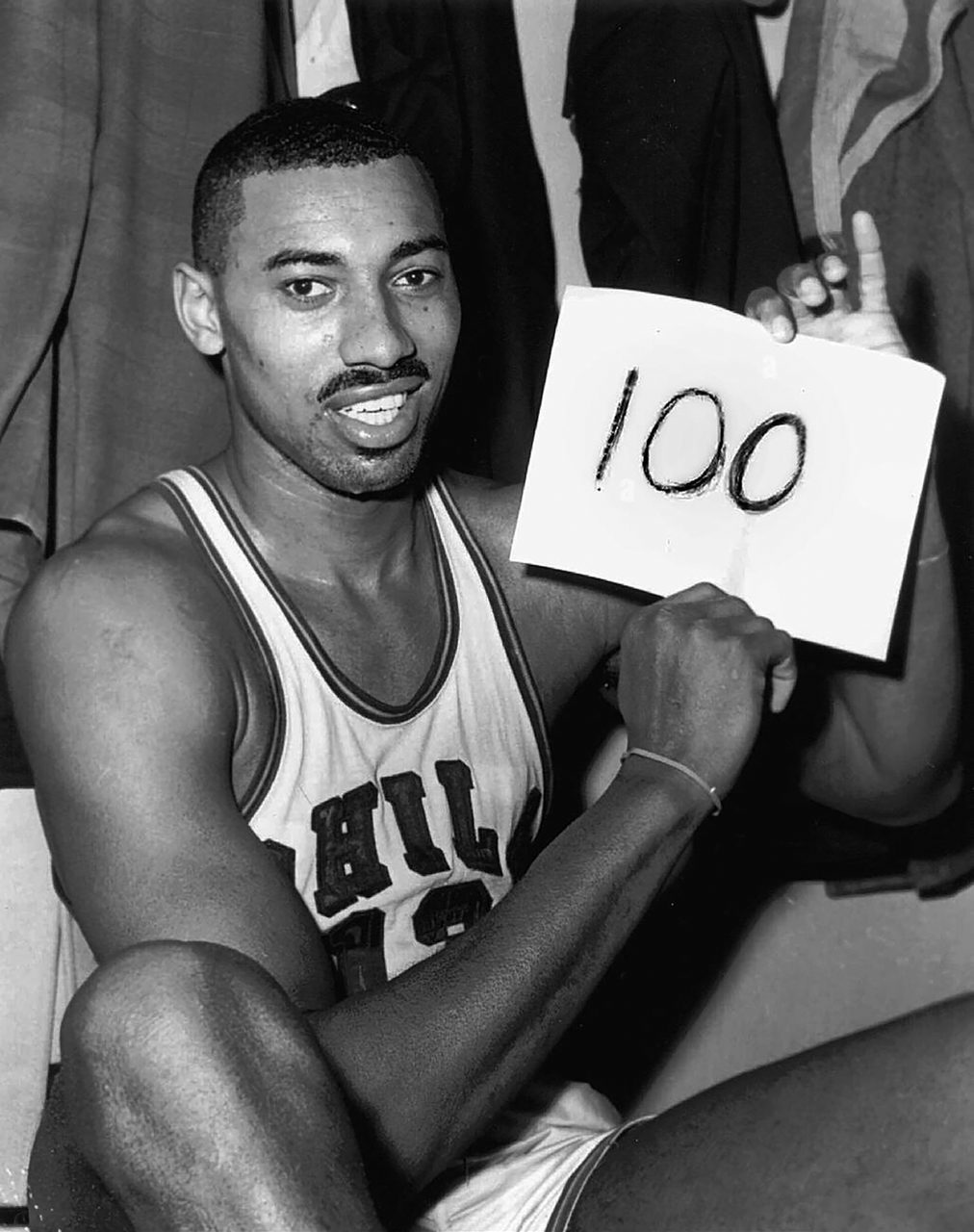 A 1962 press photograph of the basketball player Wilt Chamberlain in a dressing room holding a sign reading 100