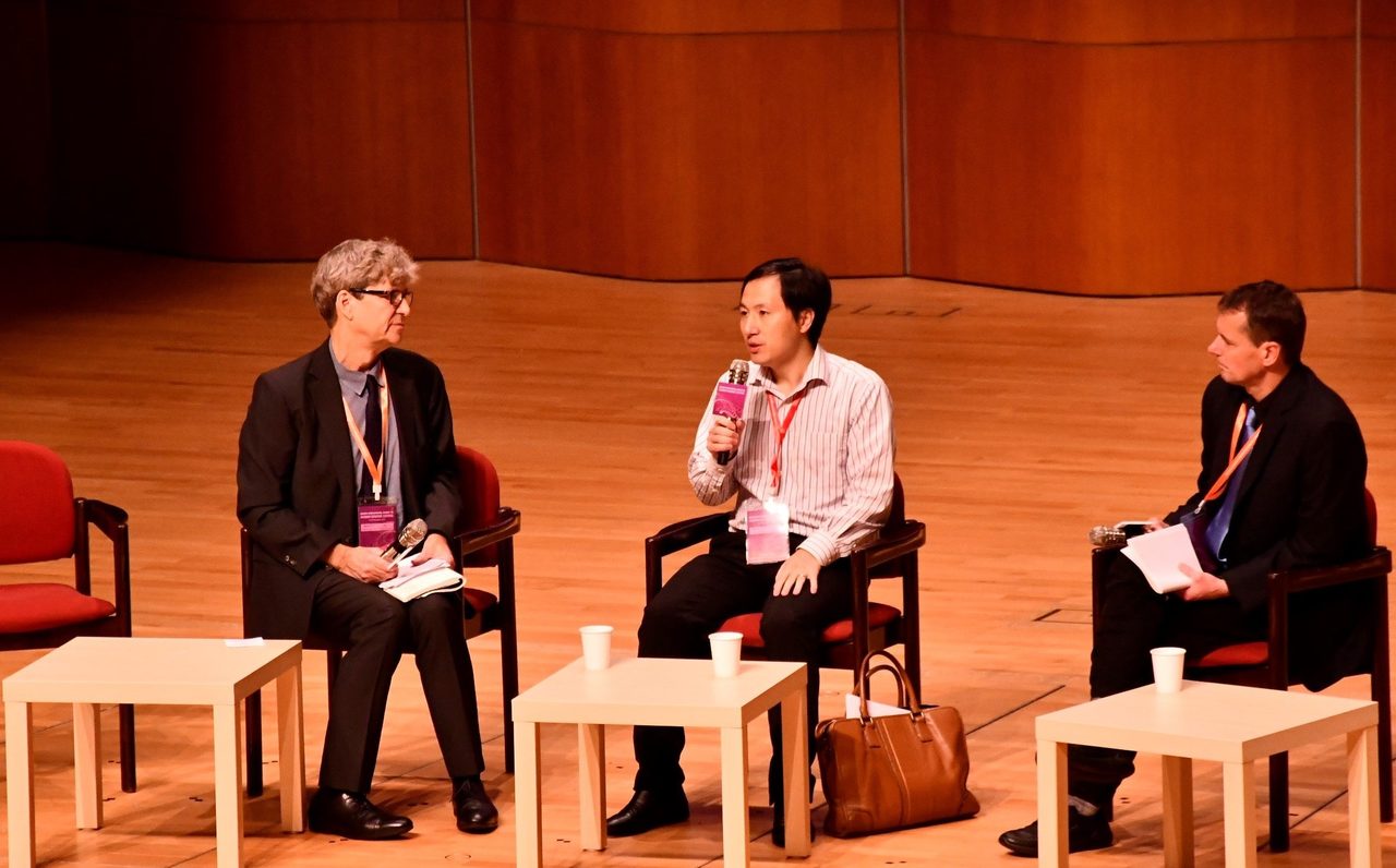 A photograph of three men seated in separate chairs on a stage at the Second International Summit on Human Genome Editing in Hong Kong on 28 November 2018
