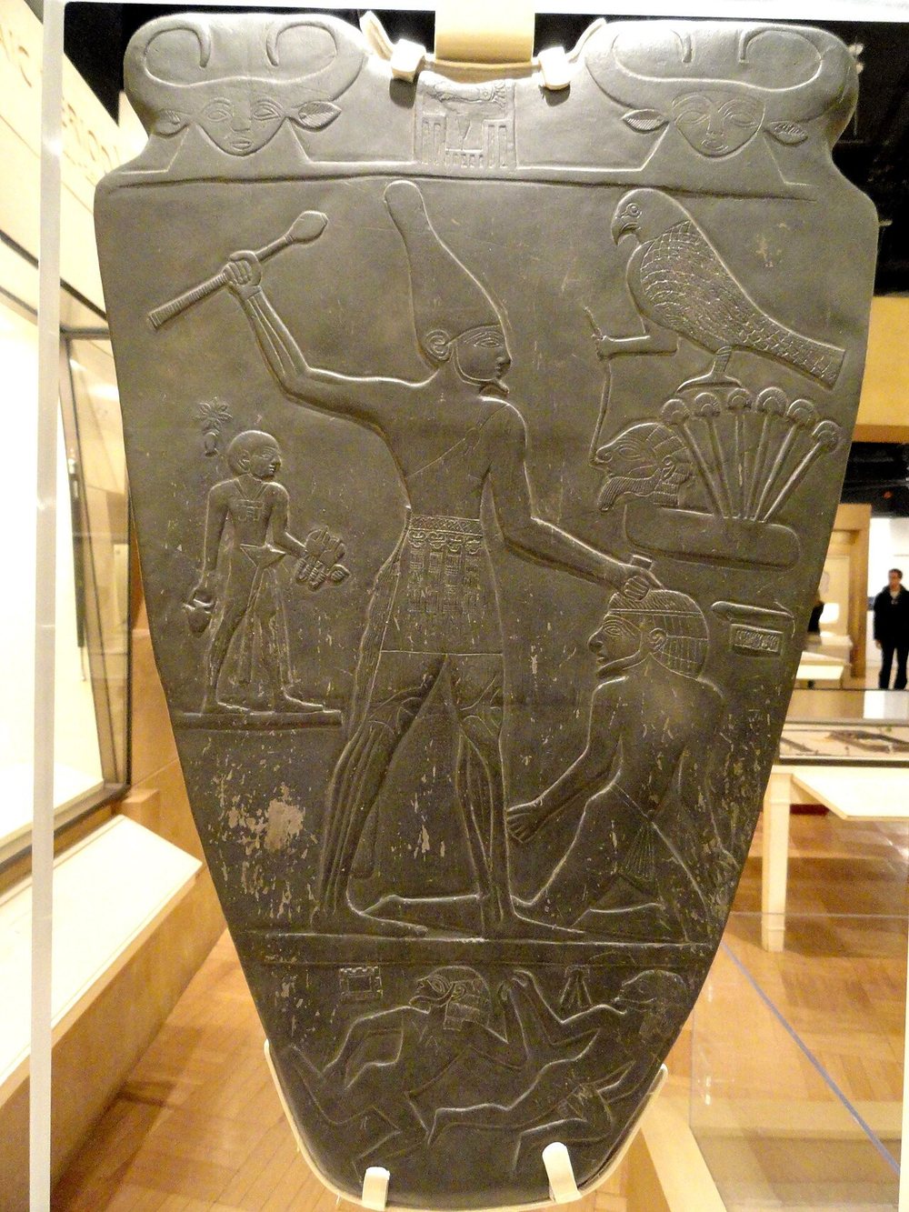 A dark shield-shaped carved stone palette displayed in a museum case: a large figure in a tall conical crown raises a mace over a smaller kneeling figure he grips by the hair, with a falcon perched on plants at upper right and smaller figures and signs around the scene