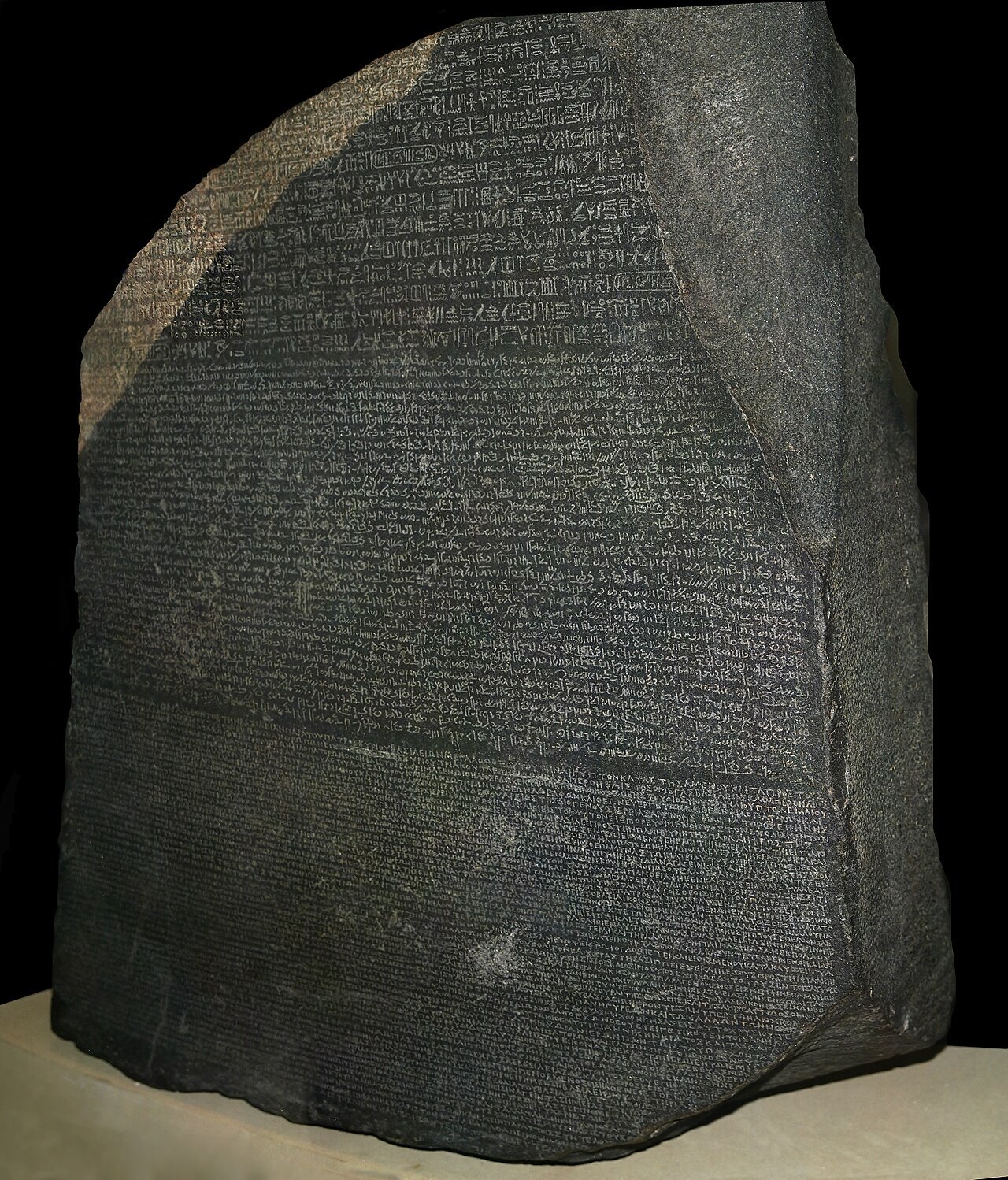 The Rosetta Stone: a large slab of dark grey granodiorite with its top corners broken away, mounted upright against a black background, its surface densely carved with three horizontal bands of different scripts, the finer lines at the bottom being Greek letters
