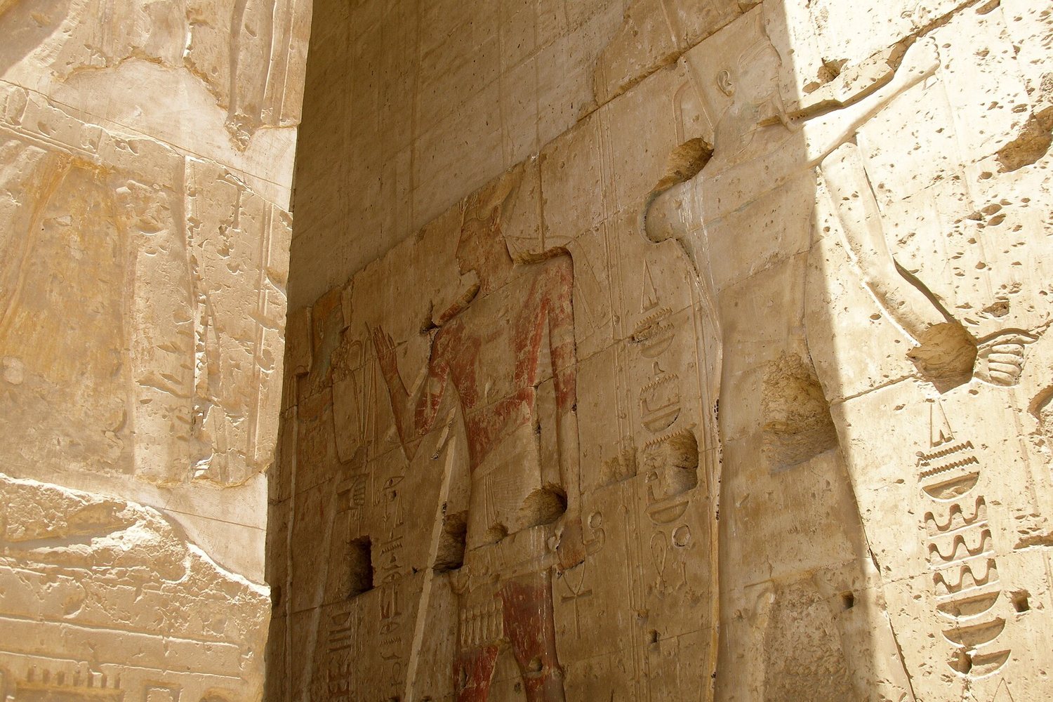 A carved and red-painted relief on a warm pale limestone temple wall in raking sunlight: a large figure striding in profile, holding a staff, flanked by columns of carved hieroglyphic signs, the surface weathered and chipped in places