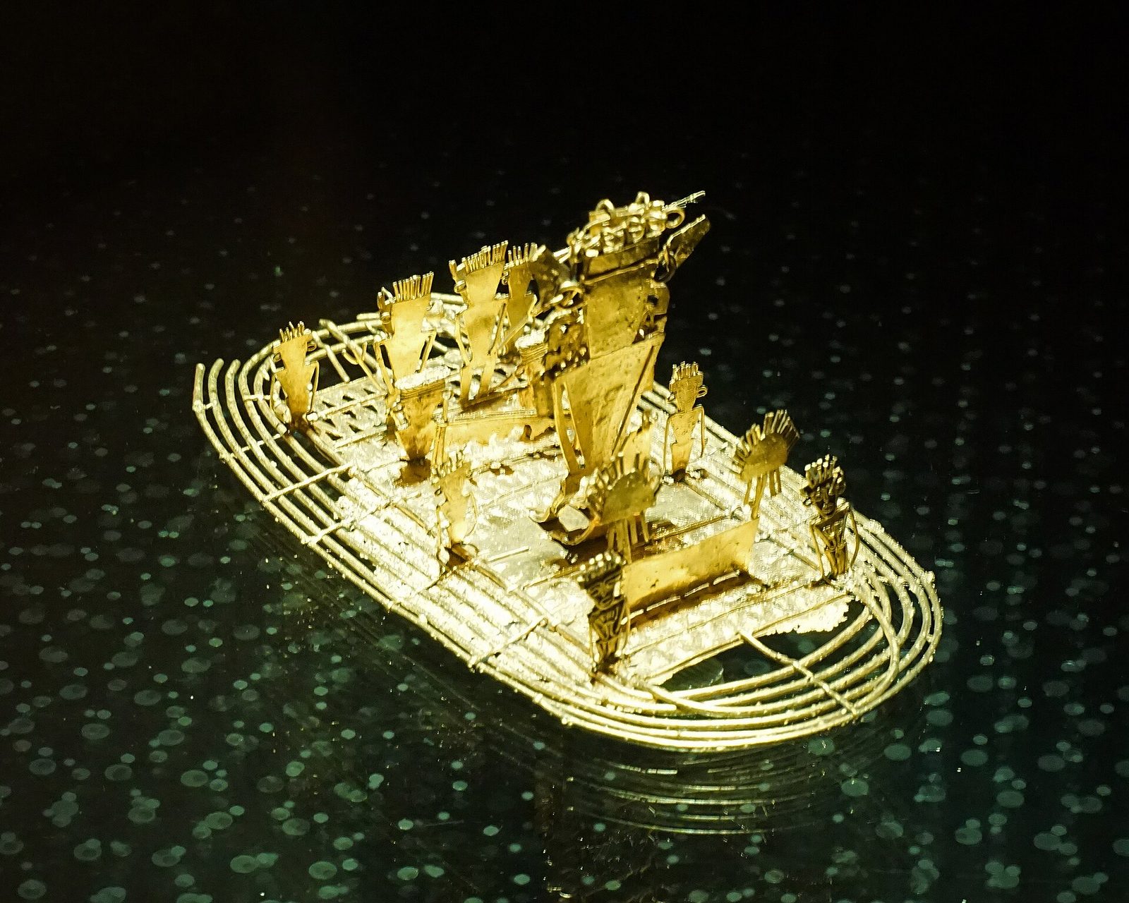 A small gold raft photographed close on a dark reflective surface. The hull is built from parallel gold wires bound into an oval platform with an upturned prow. On a raised deck at the centre stands a tall flat figure with an elaborate crown and disc ornaments, flanked by smaller standing figures in feathered or rayed headdresses, several holding staffs or vessels. The metal is warm yellow and catches the light along every wire.