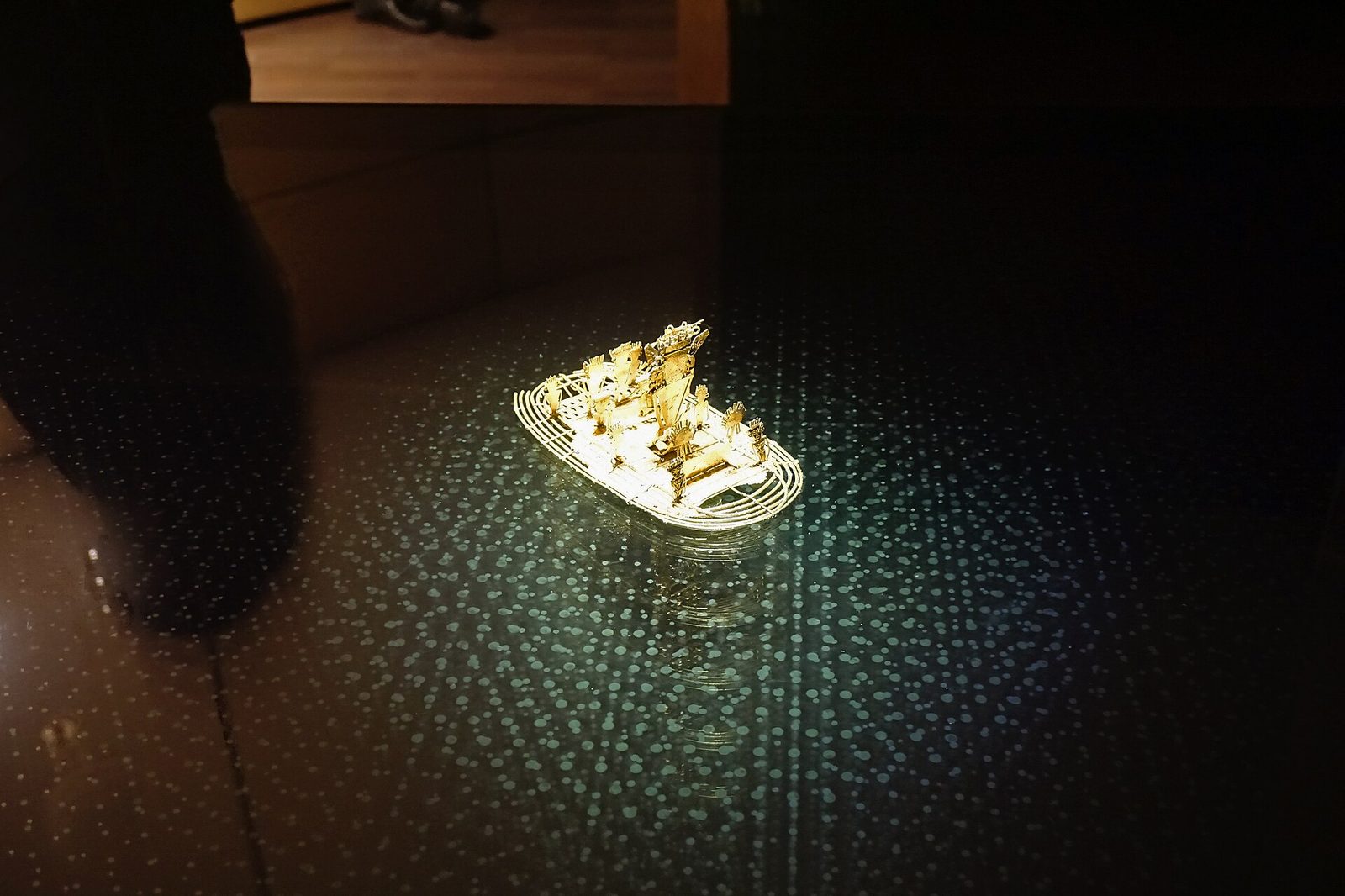 A darkened gallery. A small gold object glows alone at the centre of a wide dark plinth whose surface is scattered with faint pale points like a starfield, its reflection stretching below it. A visitor's dark silhouette blurs at the left edge of the frame. The object occupies only a small fraction of the picture.