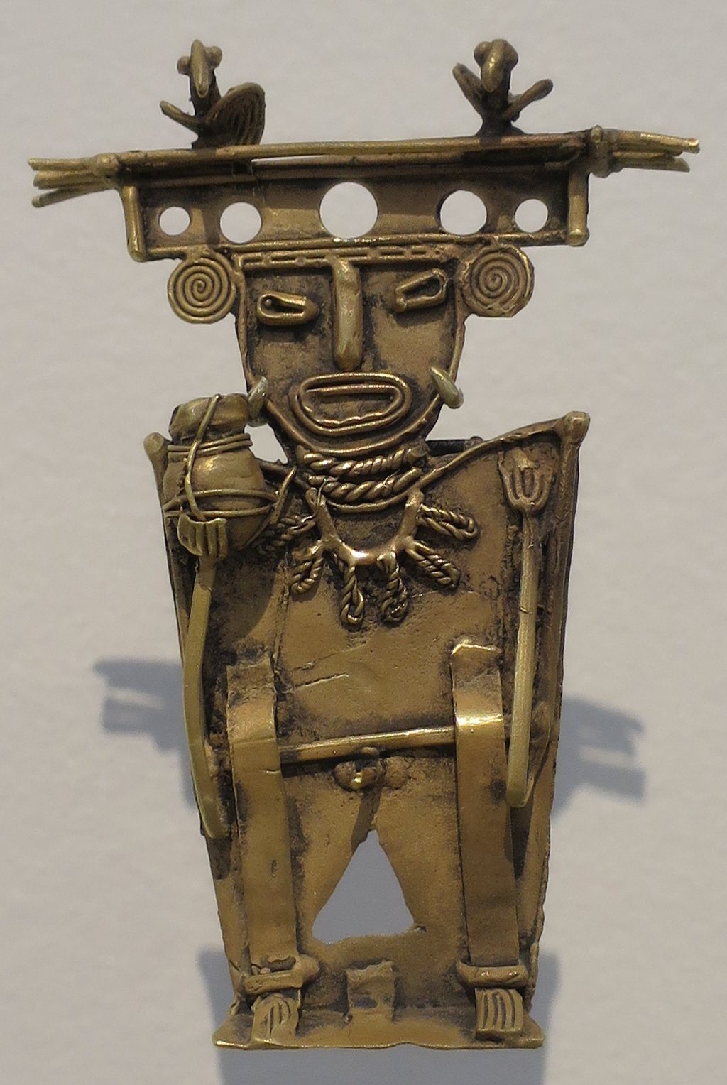 A flat cast-gold figure standing against a plain pale ground. A wide horizontal bar forms the headdress, carrying two small bird shapes at its ends and pierced by a row of round holes. The face is schematic, with spiral ear ornaments, slit eyes and an oval mouth. Rope-like collars hang at the throat, and the figure holds a staff in one hand and a small round vessel in the other.