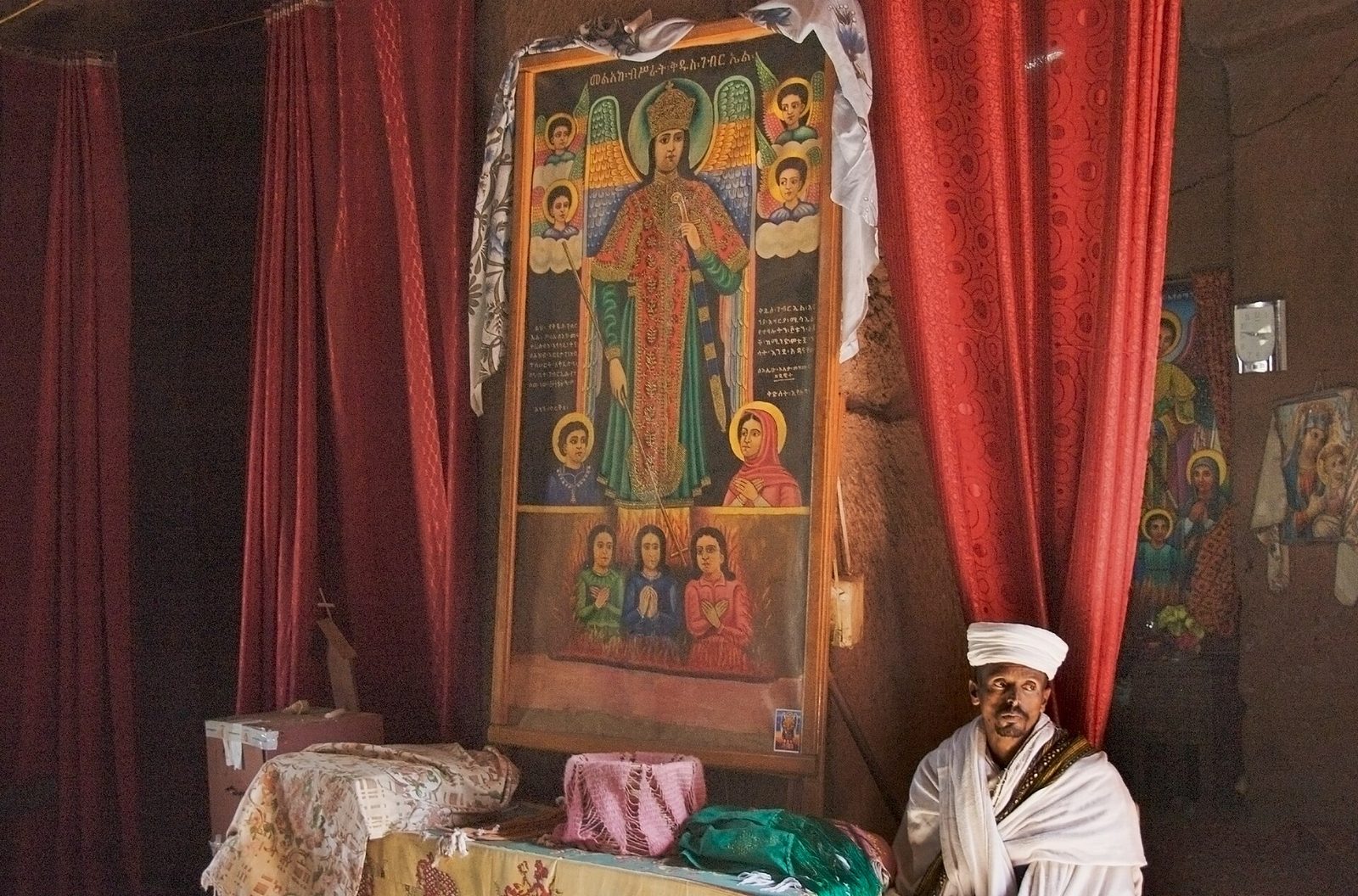 A rock-hewn church interior. A large framed painted icon shows a winged archangel in a crown and richly patterned robes, surrounded by cherub heads in clouds, with two haloed figures below and three praying figures in a lower register amid flames, and Ge'ez inscriptions on the panel. Red curtains hang on either side and a white cloth is draped over the top of the frame. A priest in white robes and turban stands at the lower right. More icons hang on the rough-hewn rock wall at the right.