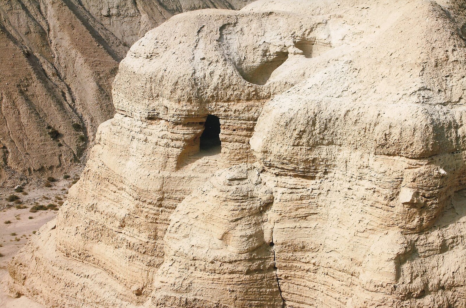 A pale, finely bedded marl cliff face with a dark rectangular opening cut into it and a second smaller opening above and to the right. Deep horizontal stratification runs across the whole face, and dry desert slopes with scattered scrub lie behind and to the left.