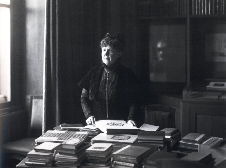 A photograph of Elisabeth Foerster-Nietzsche standing at a table covered with stacked books, both hands resting on an open volume