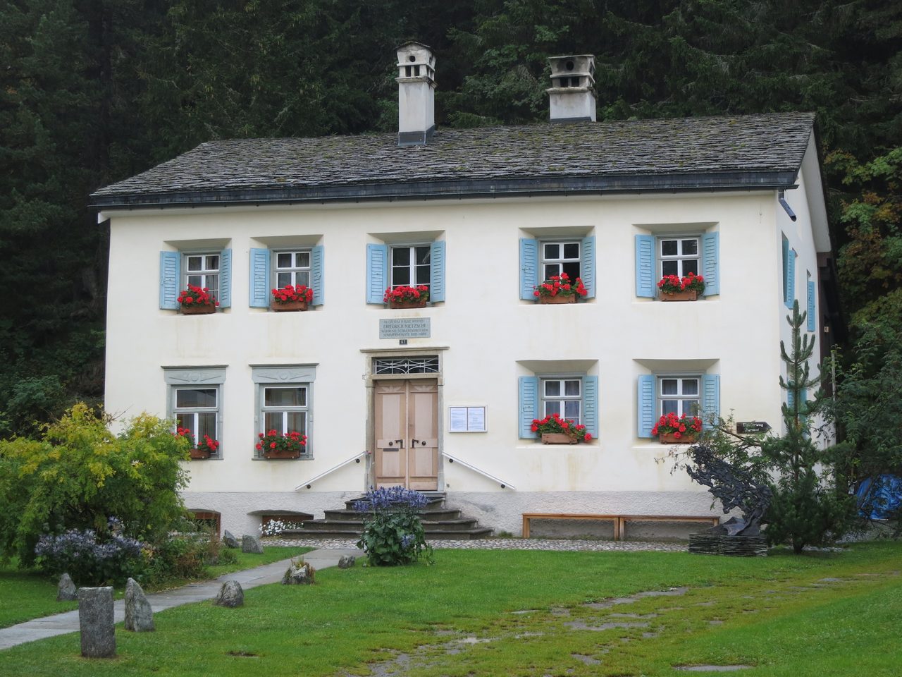 A cream alpine house with pale blue shutters and red geraniums at the windows, backed by dark forest, with an information panel beside the door