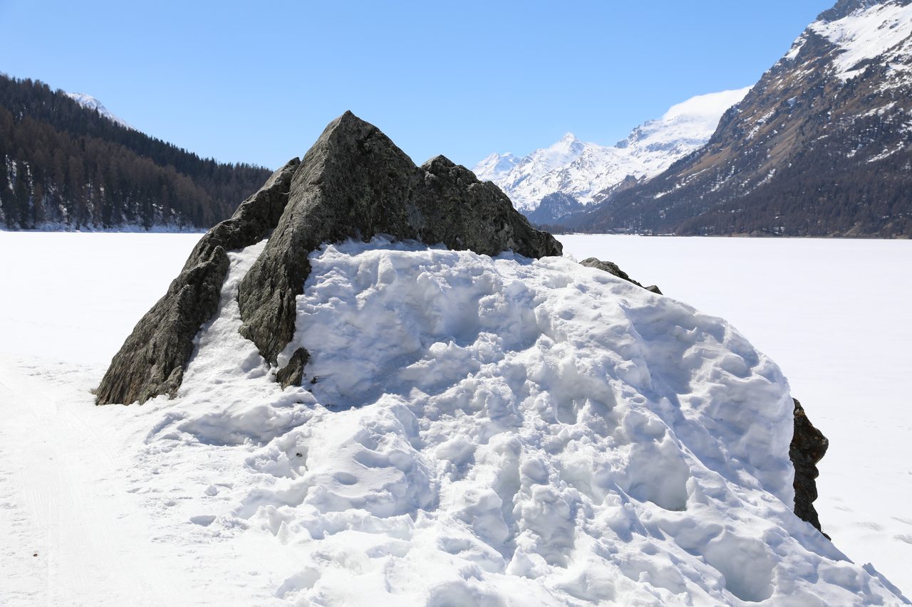 A pointed grey rock rising out of deep snow beside a frozen lake, with forested slopes and snow-covered peaks behind it, a cloud cap over one summit