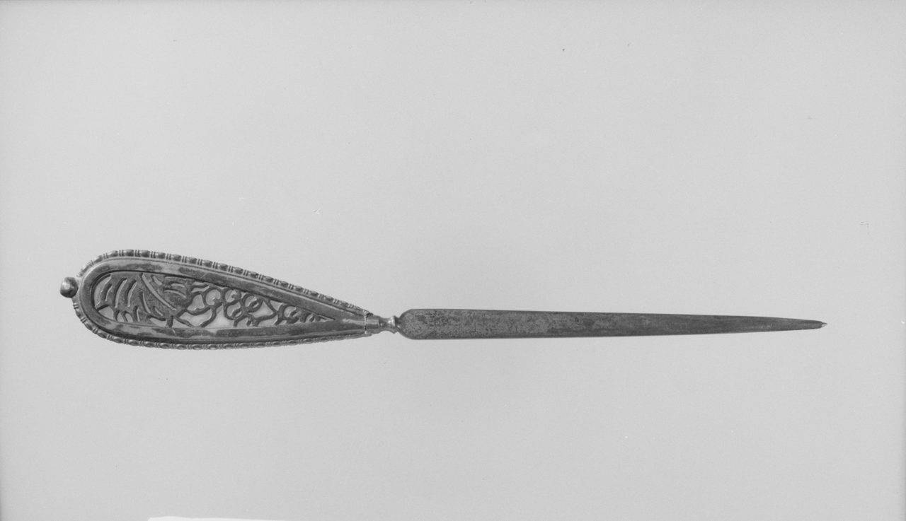 A documentation photograph of a metal letter opener made by Tiffany Studios, lying on a plain grey ground with a long tapering blade and a broad openwork teardrop handle