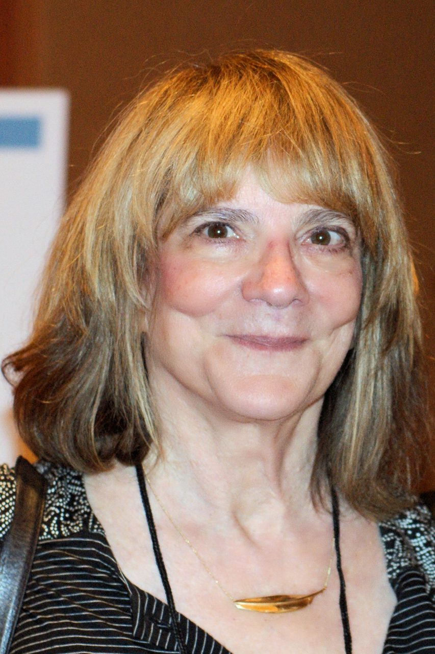 A close colour portrait photograph of a woman with shoulder-length fair hair and a full fringe, looking just past the camera with a slight smile, wearing a dark patterned top with a black cord and a fine gold chain carrying a curved gold bar pendant, a dark strap crossing the shoulder at the left of the frame, against a warm brown out-of-focus background, with the pale edge of a white panel carrying a blue block at the far left of the frame