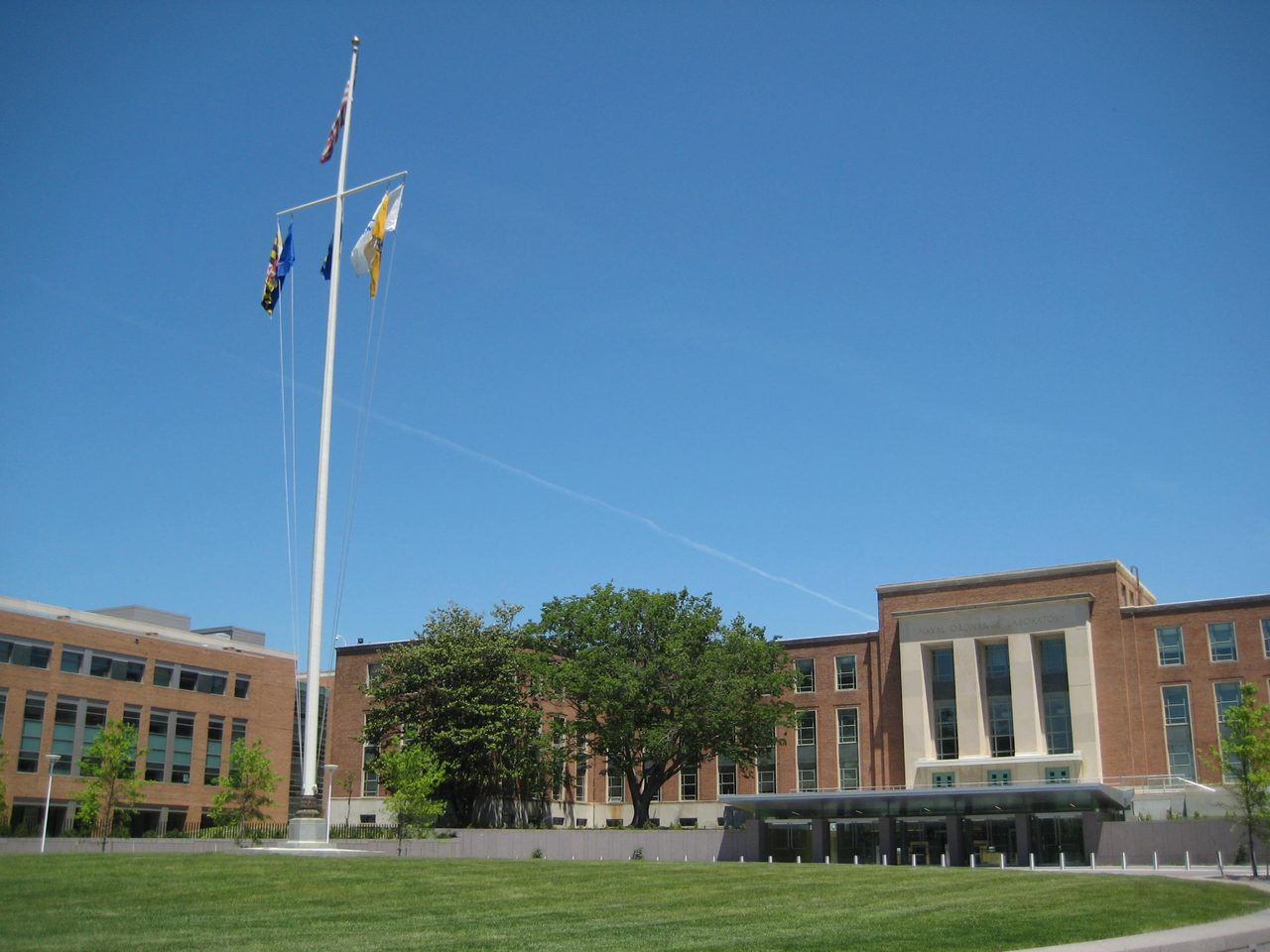 Two office buildings on a government campus, with a flag pole and lawn in the foreground under a blue sky