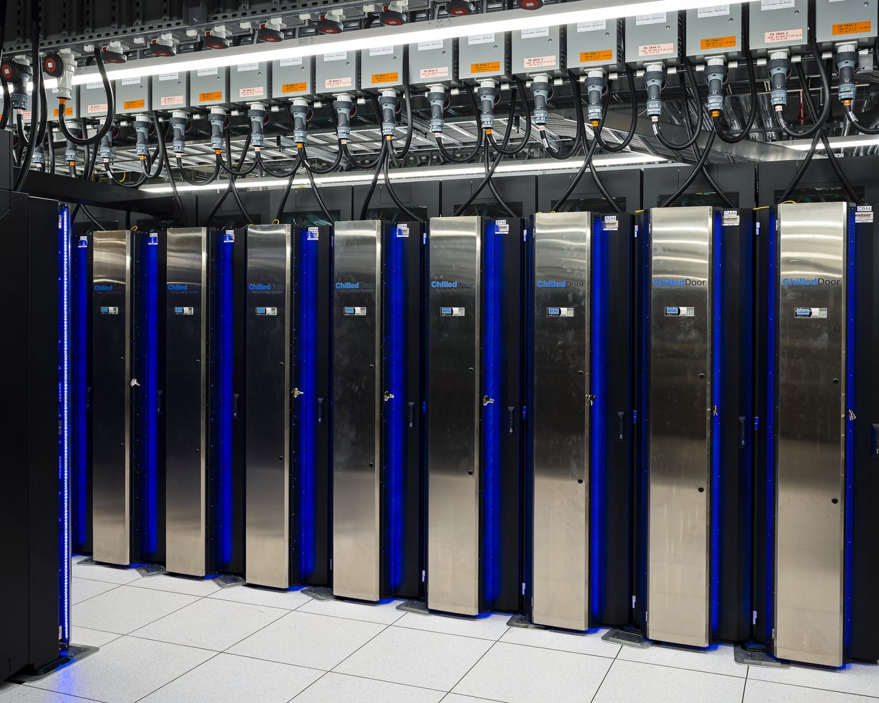 A row of tall black server cabinets with brushed stainless-steel doors and blue edge lighting, standing on a white raised floor beneath an overhead tray of black cables and labelled power connectors: the Frontier supercomputer installed at Oak Ridge National Laboratory, photographed in July 2022