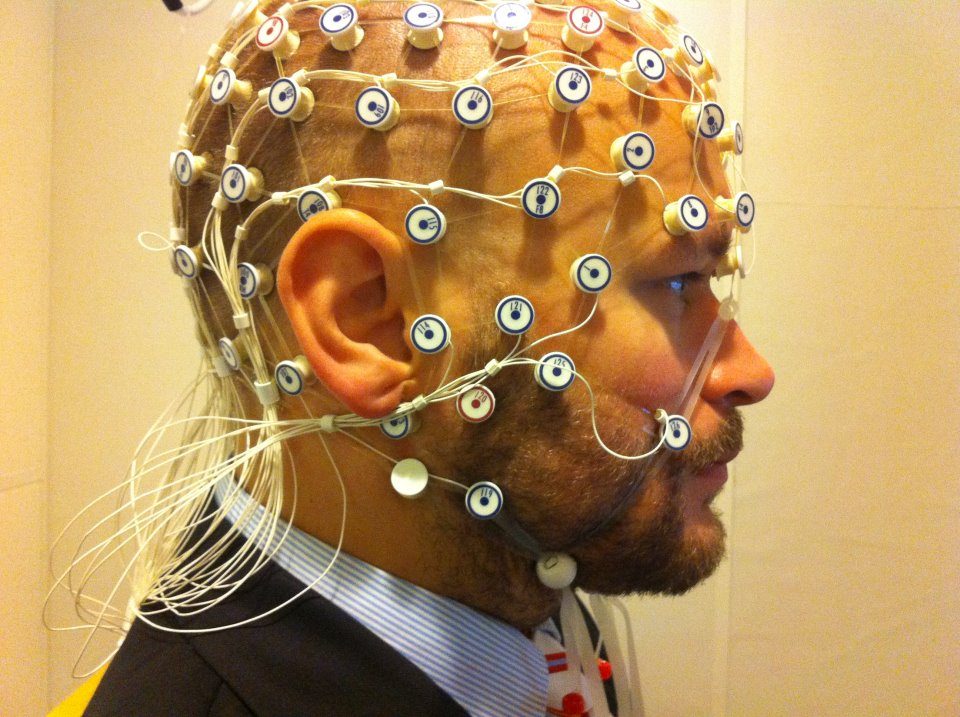 A close profile photograph of a bearded man wearing a dense net of small round electrodes over his scalp and face, with a thick bundle of pale cables running back from his head