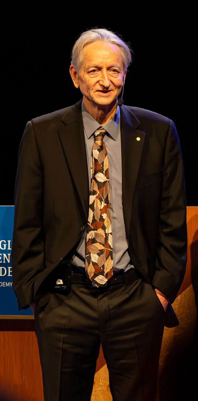 A full-length photograph of Geoffrey Hinton standing on a stage wearing a headset microphone, with an academy banner visible behind him