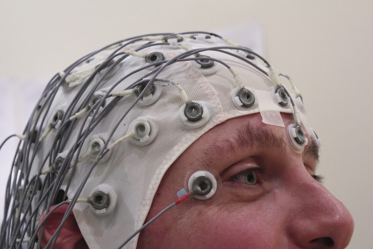 A close photograph of a man's head in profile wearing a white mesh EEG cap studded with ring electrodes and trailing grey cables, one electrode taped near his eyebrow