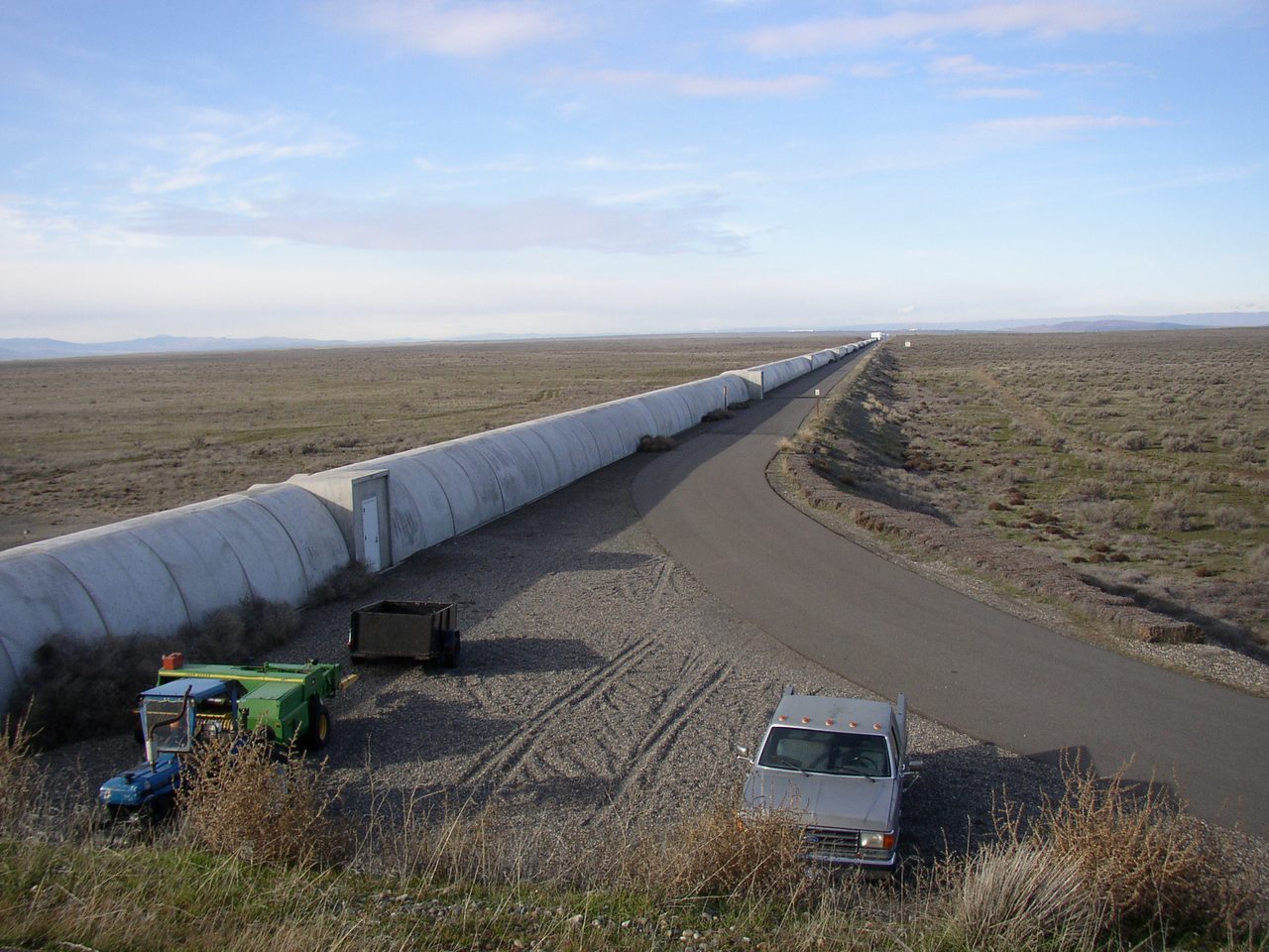 A ground-level photograph looking along a long, low, straight enclosed tube that runs away from the camera across flat open country toward the horizon