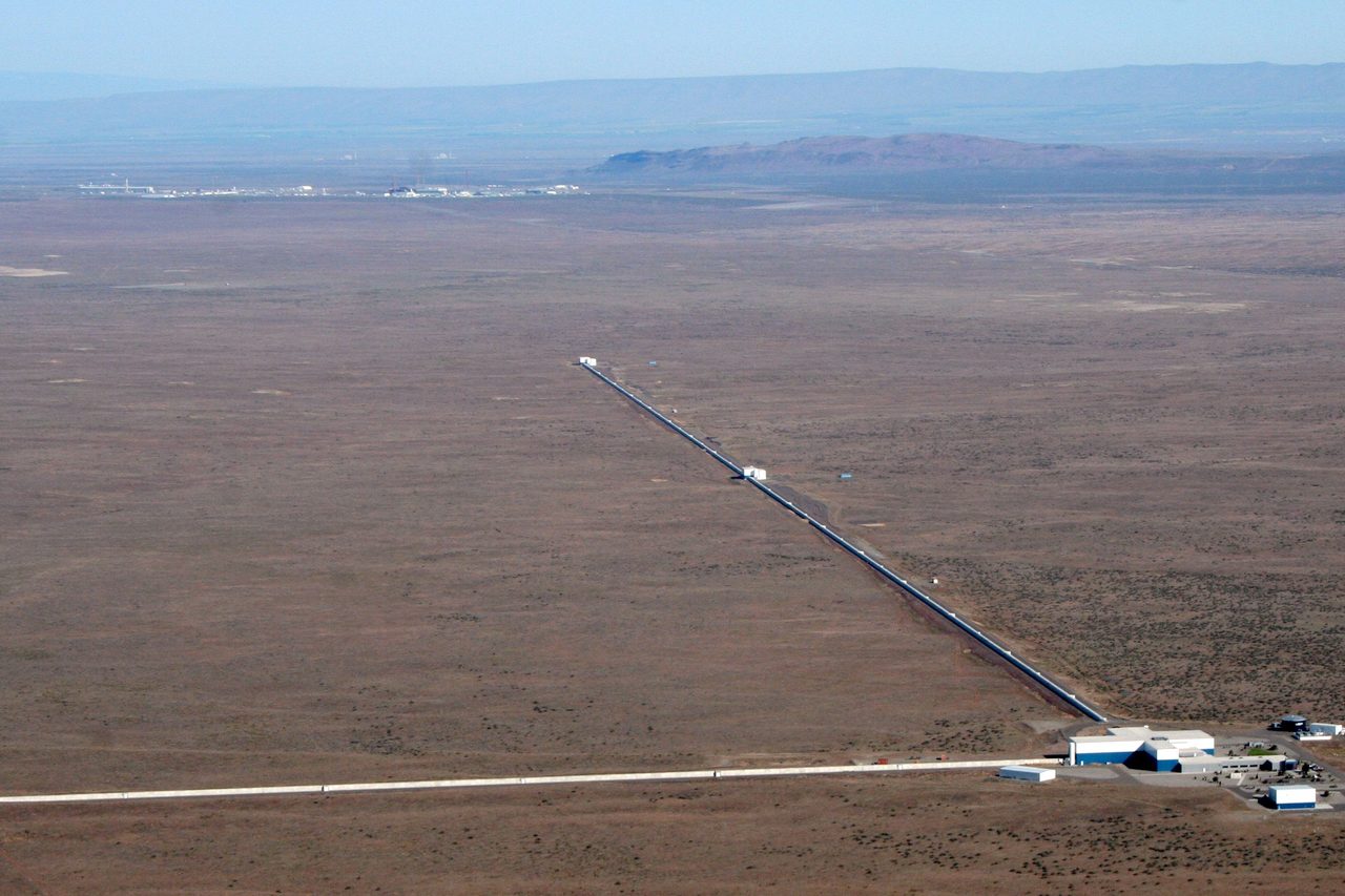 An aerial photograph of a scientific facility on flat open ground: a low cluster of buildings at the corner of two long straight enclosed tubes running away at a right angle toward the horizon