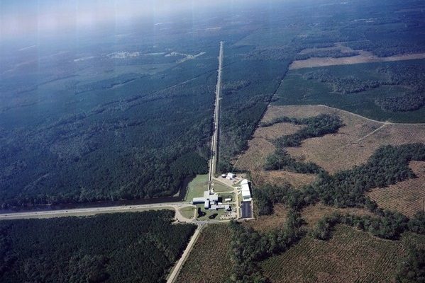 An aerial photograph of a facility in flat wooded country: a cluster of buildings with one long straight enclosed tube running away from it toward the horizon, and a public highway crossing the frame at an angle to it