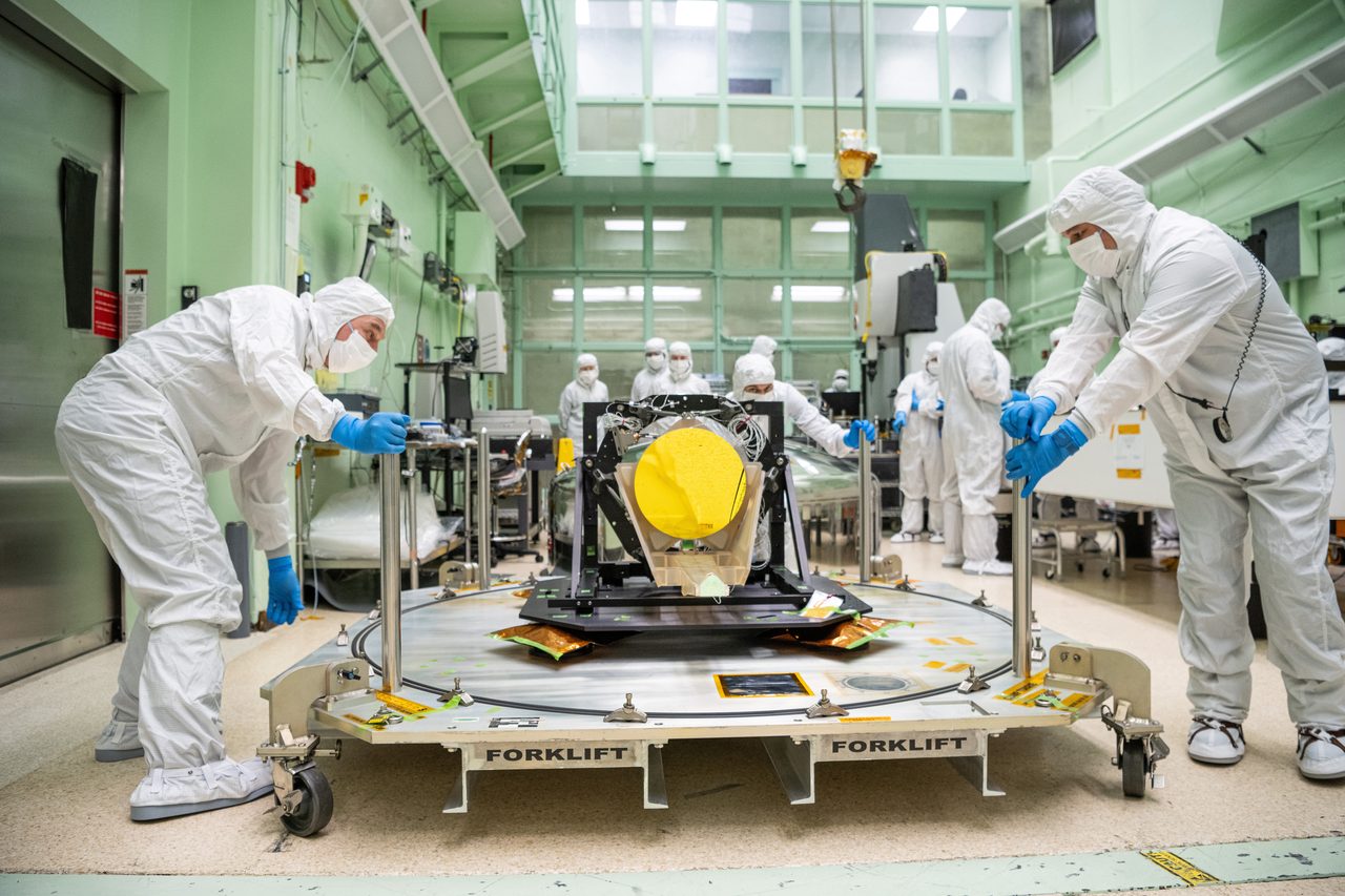 A photograph inside a cleanroom: technicians in protective coveralls stand around a black open frame holding a tapering optical barrel with a large gold-coloured disc at its centre, mounted on a wheeled pallet