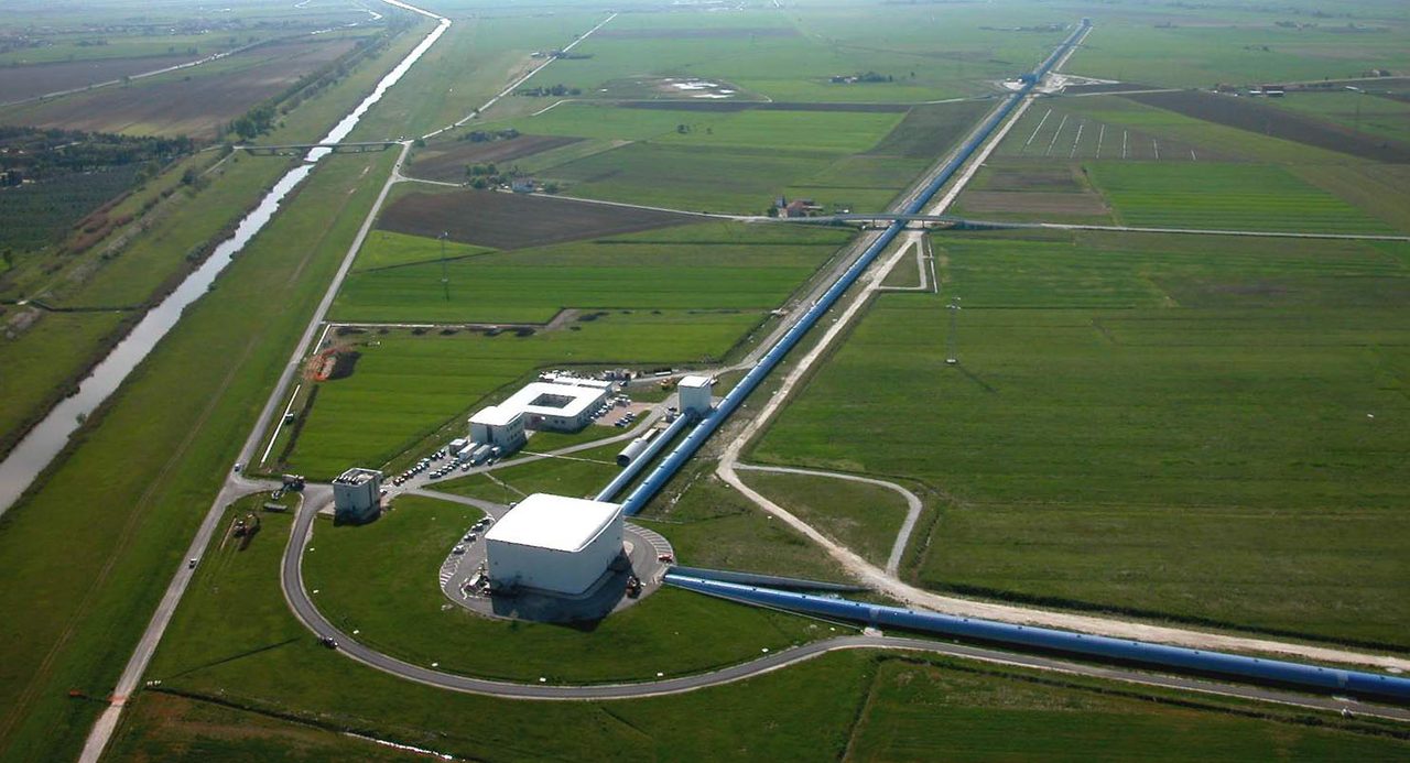 An aerial photograph of a research site in cultivated flat country, showing clustered low buildings with one long straight enclosed tube running away from them across the fields toward the horizon and a second running out of the frame in the foreground at an angle to it
