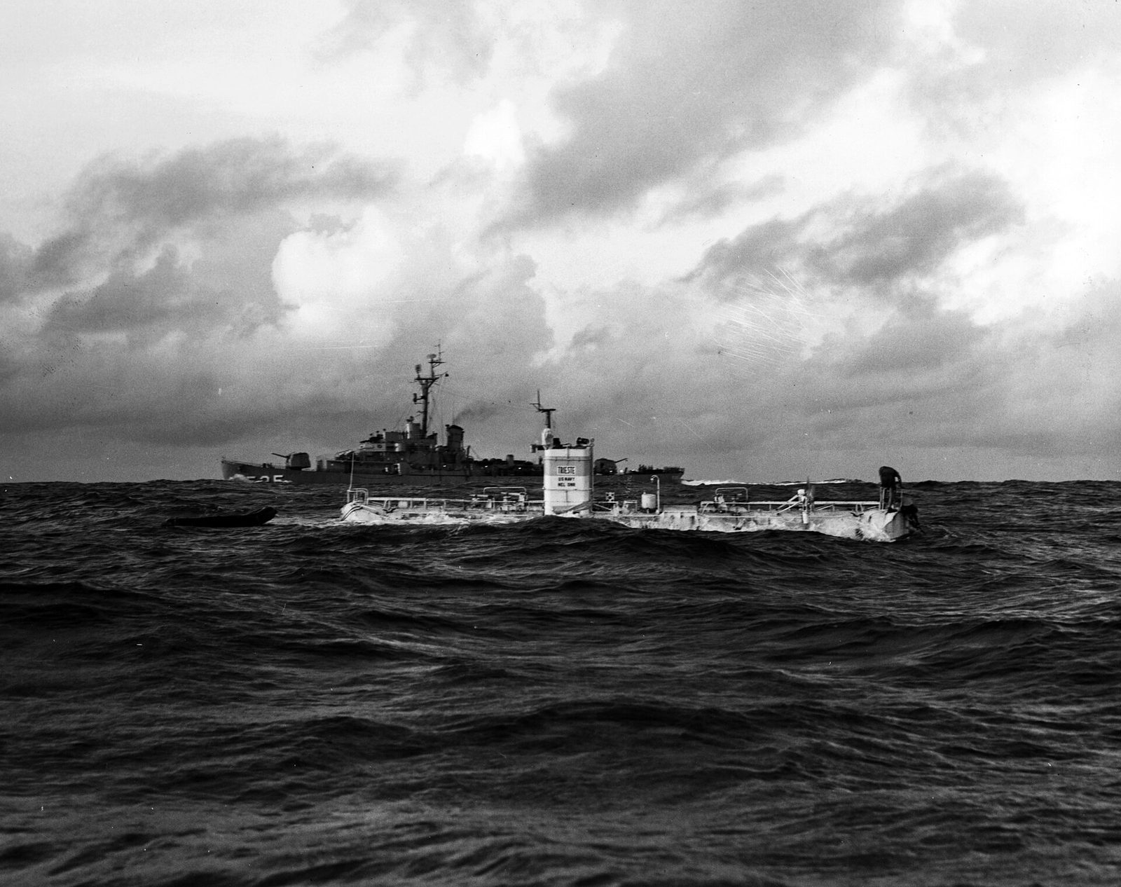 A black and white photograph. The bathyscaphe Trieste floats low in a choppy grey sea, its long flat float hull almost awash, with a white conning tower amidships lettered TRIESTE and U.S. NAVY and railings along the deck. A small dark inflatable sits at the left. A US Navy destroyer escort stands off in the middle distance. Heavy cloud fills the upper half of the frame.