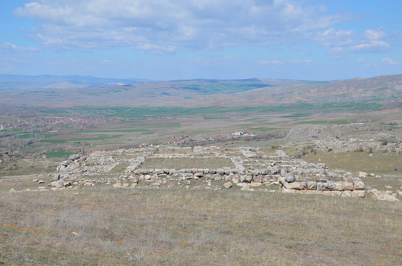 Stone wall foundations one or two courses high, laid out in rectangular room plans on dry yellow-brown grass in the foreground. Beyond them a wide plain: green cultivated strips, pale ploughed fields, a modern village with red roofs and a white minaret, low bare hills running to the horizon, and a big sky with scattered cloud.