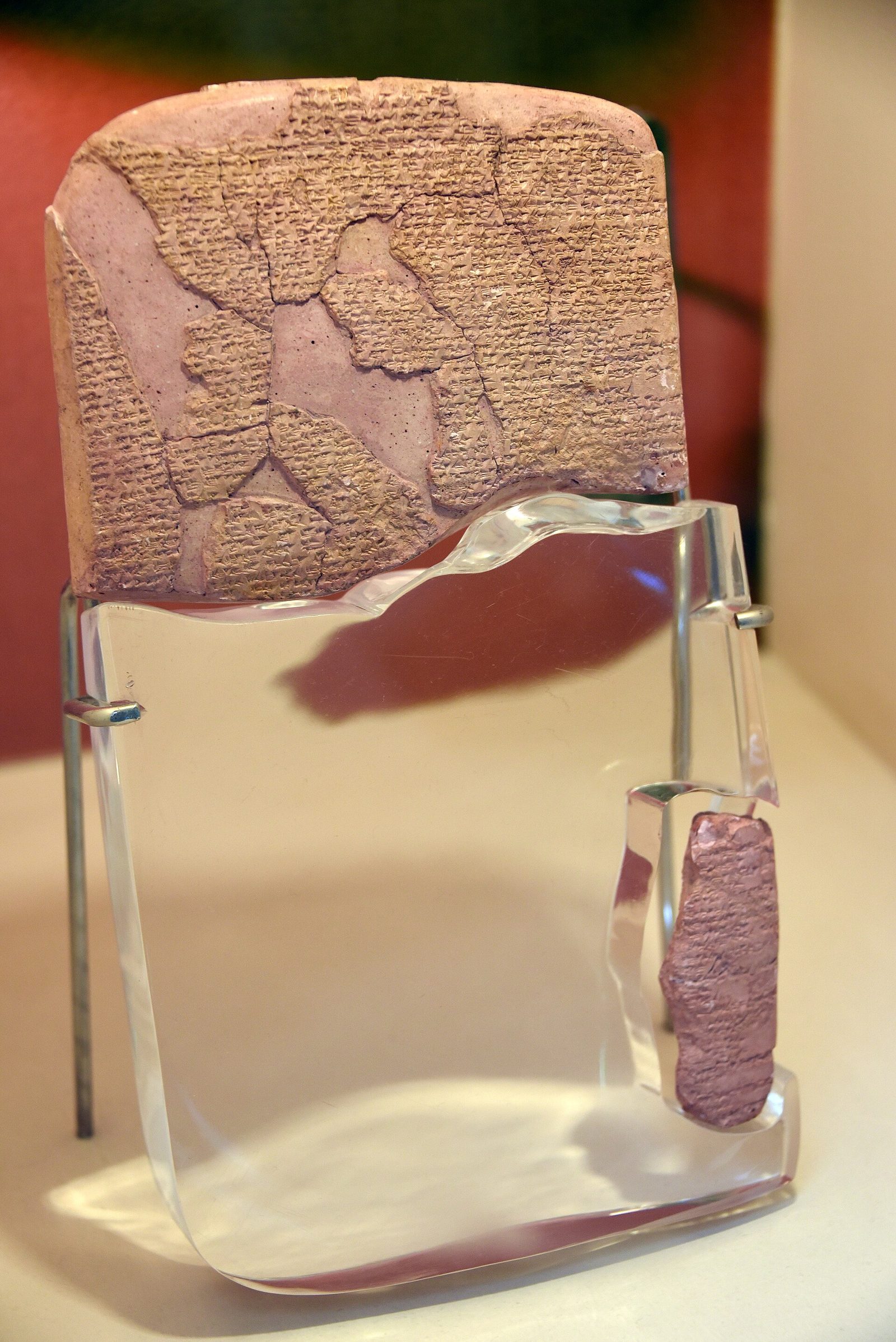 Only the upper portion of a clay tablet survives, densely covered in cuneiform and cracked into many joined fragments with pale plaster fills between them. It is mounted on a clear perspex support cut to the outline of the missing part, which occupies the lower two thirds of the object, held by metal clips on two thin rods. A single small separate fragment is clipped to the perspex low at the right, isolated in the empty space. Warm gallery light, a pink and red wall behind, a cream plinth.