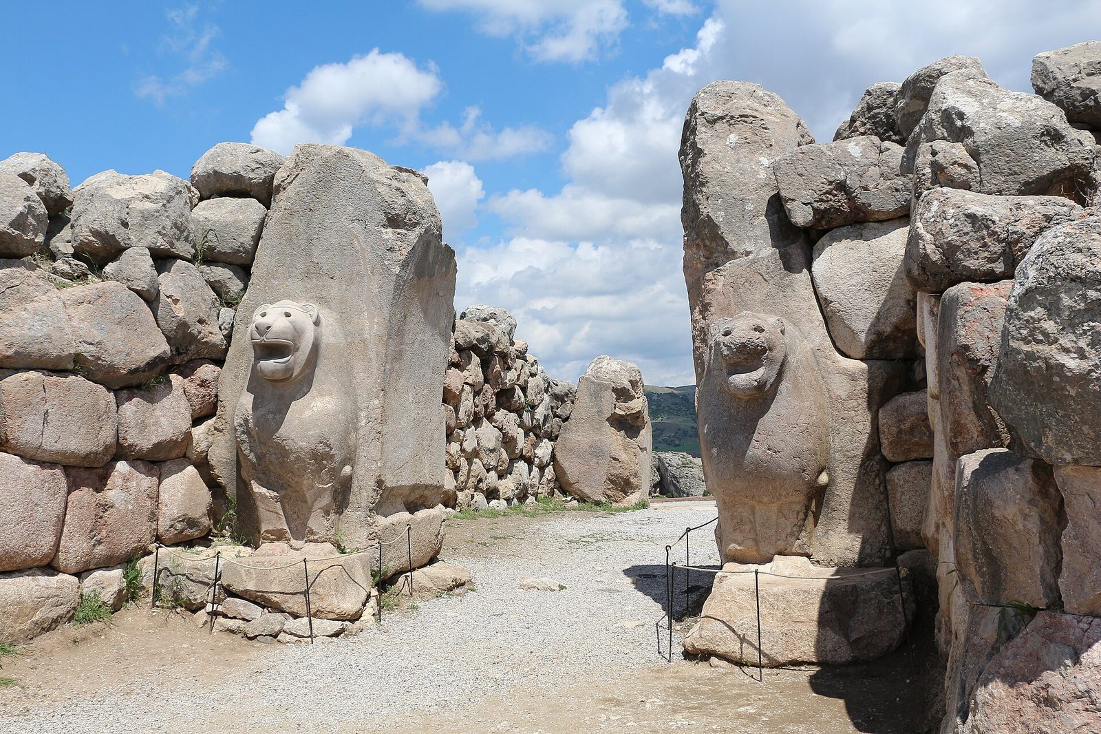 Two lions carved in relief on massive stone blocks, facing each other across a gravel passage. The left lion is sharp and legible with a smooth muzzle, defined ears and an open mouth. The right lion is deeply eroded, its face pitted and its features blurred. Cyclopean walls of rough boulders run away on both sides, thin modern metal railings on low posts stand around both bases, and green hills and a blue sky with cumulus show through the gap.