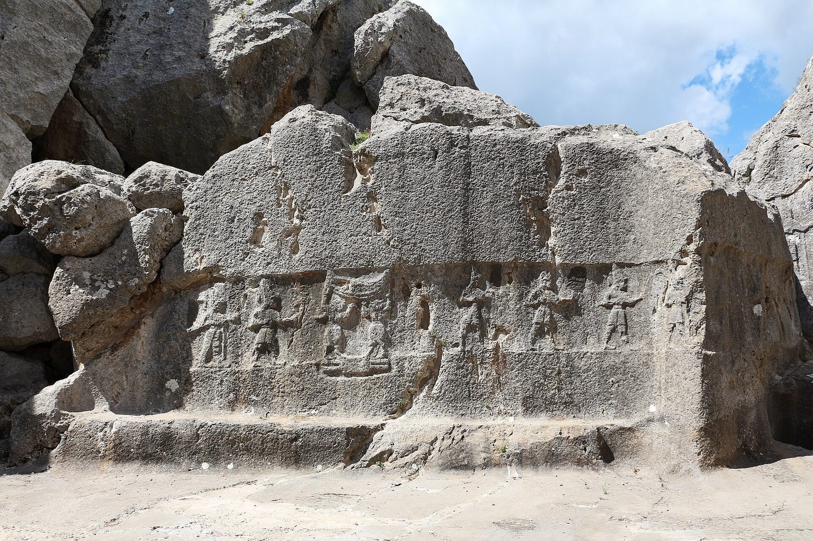 A natural limestone rock face, weathered and cracked, carrying a horizontal band of low relief: a procession of standing figures in profile wearing tall pointed caps, arms extended forward. The relief is badly worn, the rock above the band is blank and pitted, several figures are reduced to outlines, and a large area at the right has lost its carved surface entirely. Bare pale rock underfoot, blue sky and cloud at the upper right.