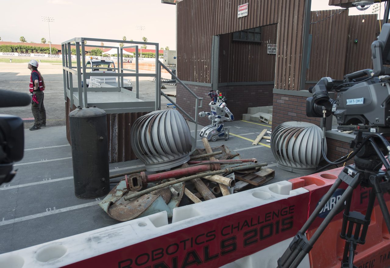 A humanoid robot on a competition course approaching a low obstacle, with an official in a hard hat at the left, and television cameras and event barriers around the course