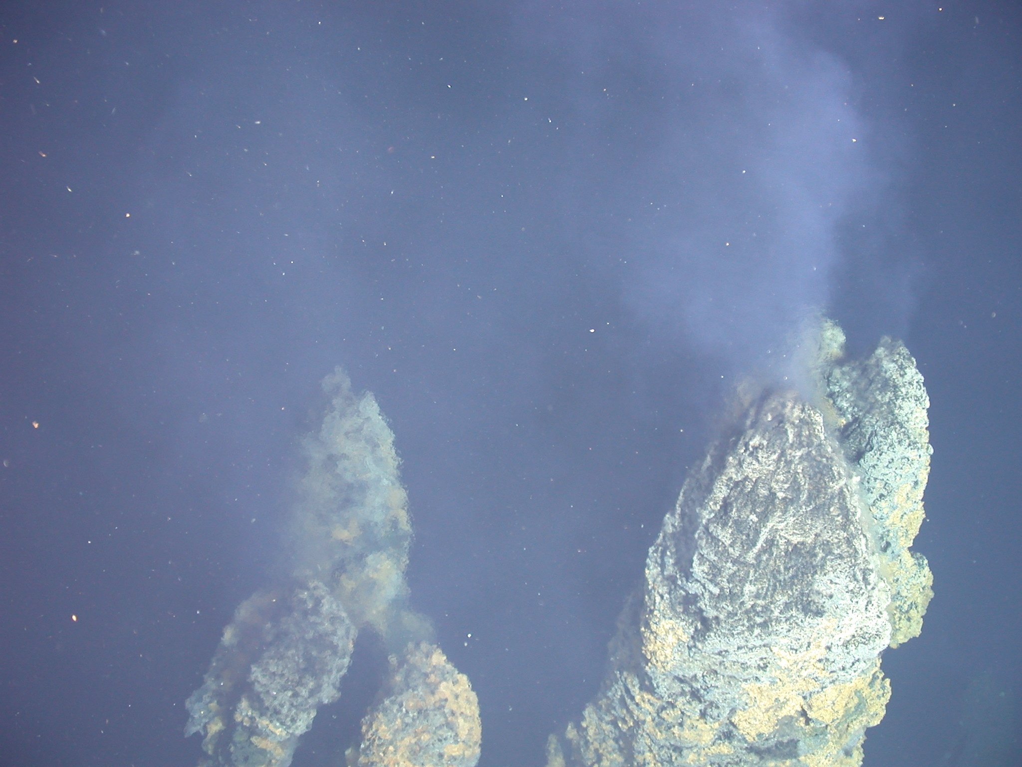 Two tall hydrothermal vent chimneys encrusted in yellow, white, and blue-grey mineral deposits rise from the seafloor, each venting a shimmering, particle-laden plume upward into dark blue-black deep-sea water